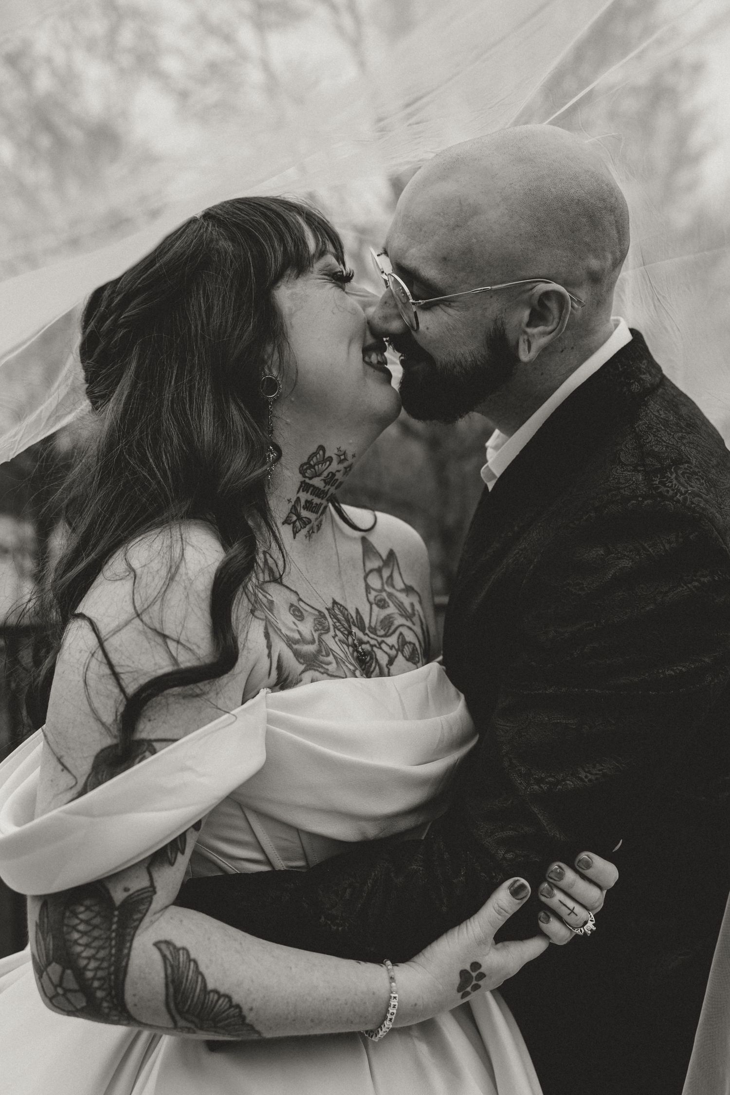 Black and white photo of tattooed bride and bald groom laughing under veil on wedding day.