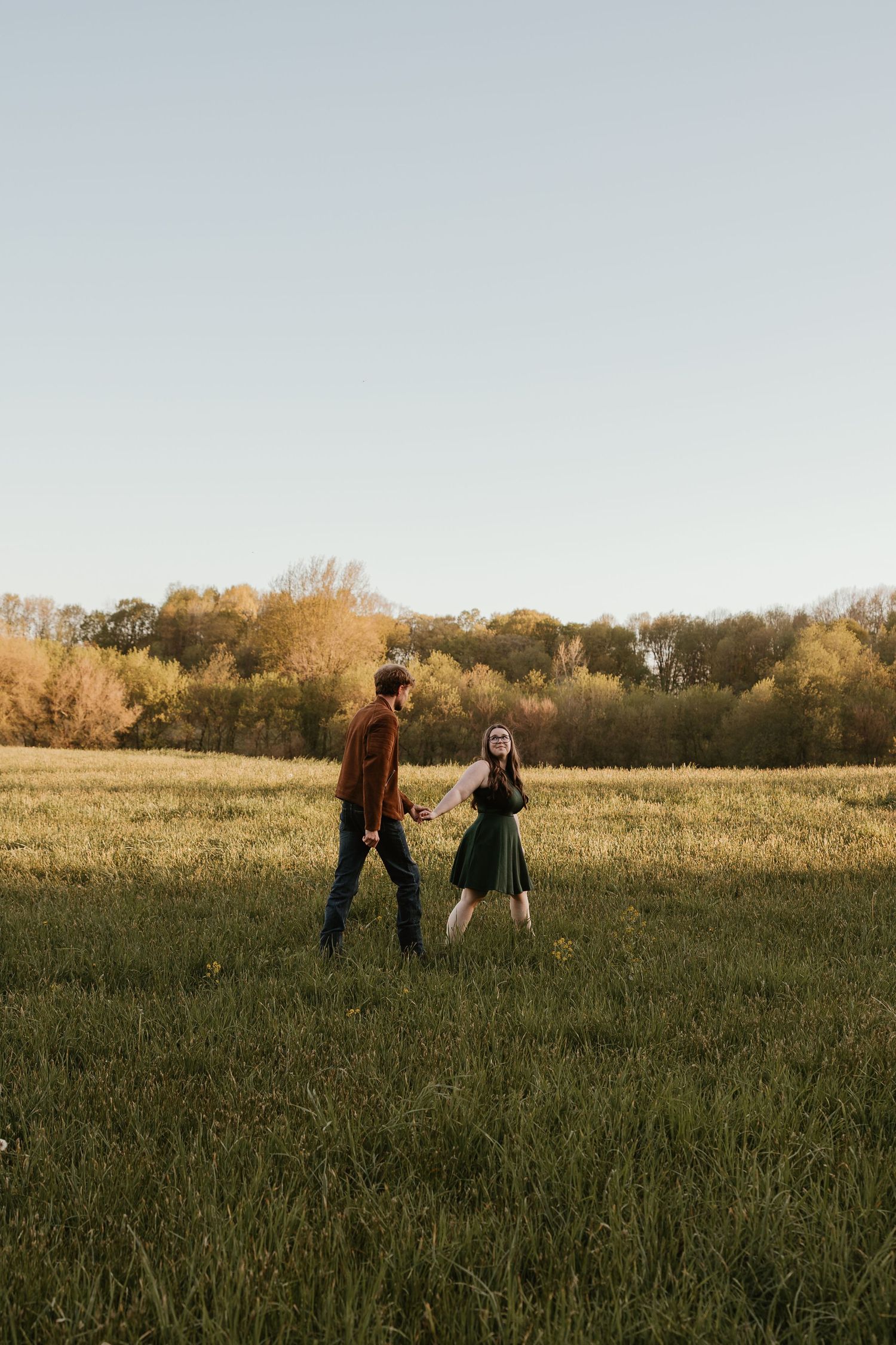 A couple walks hand in hand through a green meadow with autumn trees in the background during golden hour.