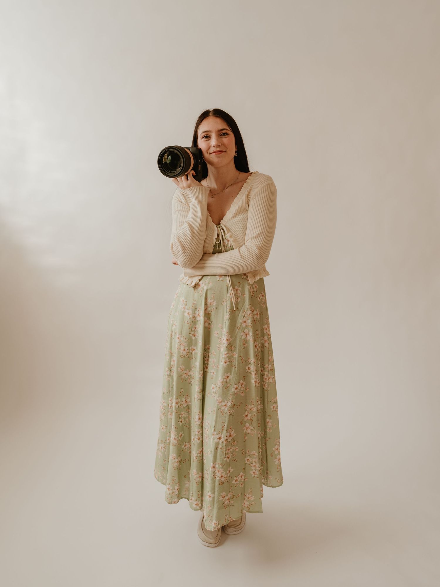 Woman in floral maxi skirt and cream cardigan holding a professional camera against a light background.