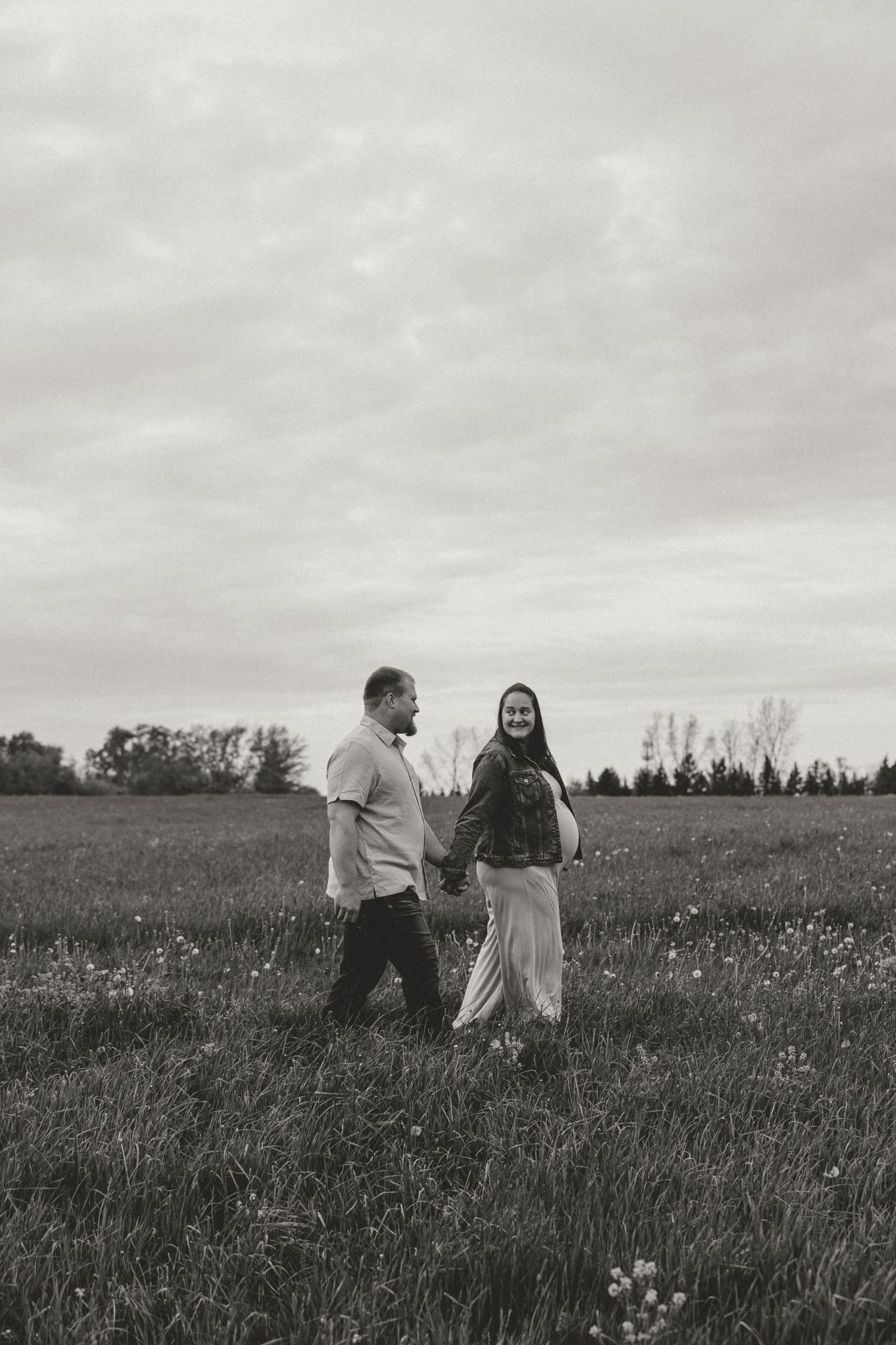 Black and white photo of a couple holding hands walking through a grassy field with trees and cloudy sky.
