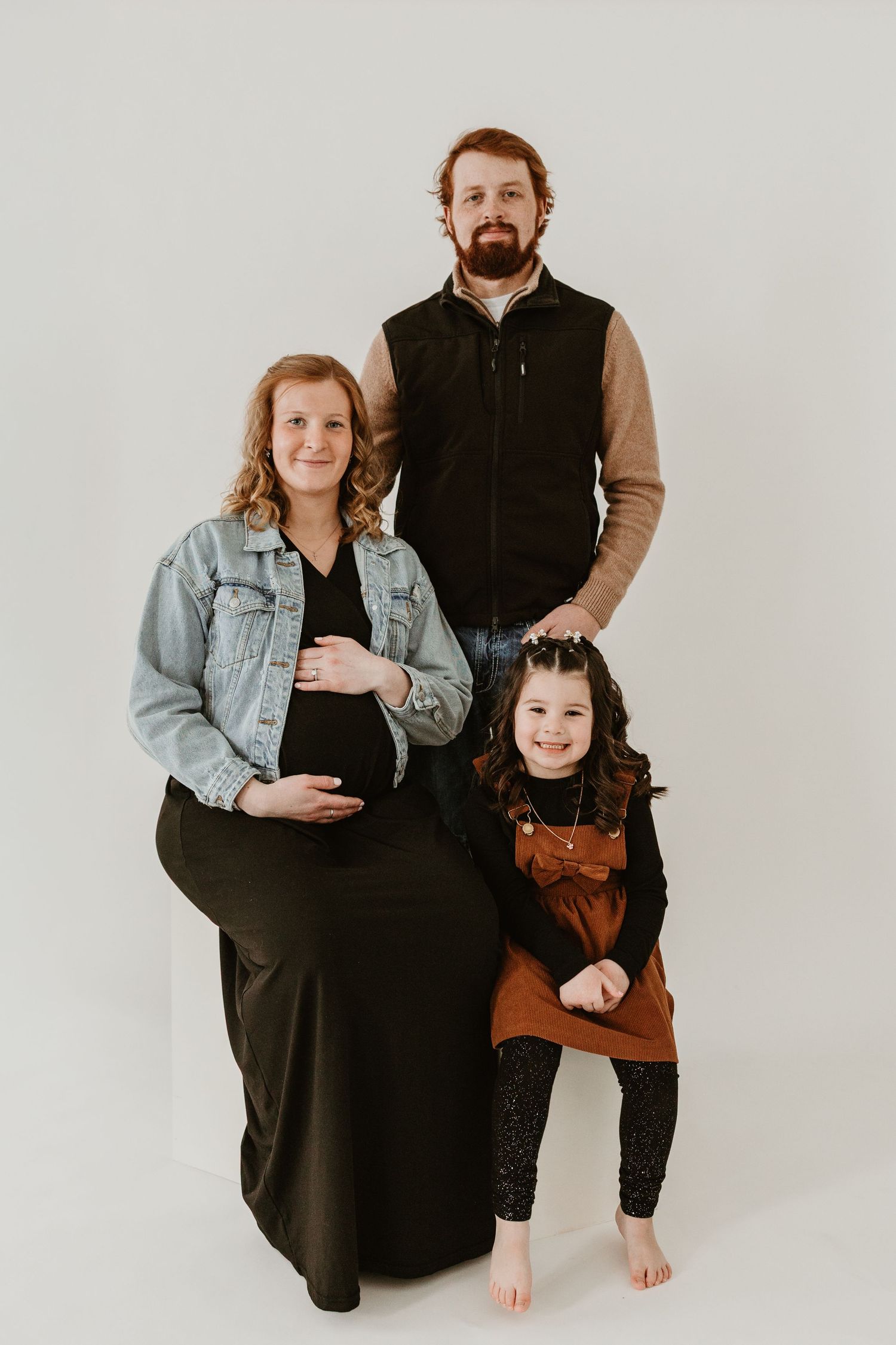 Maternity family portrait with pregnant mother, bearded father in vest, and young daughter in studio.