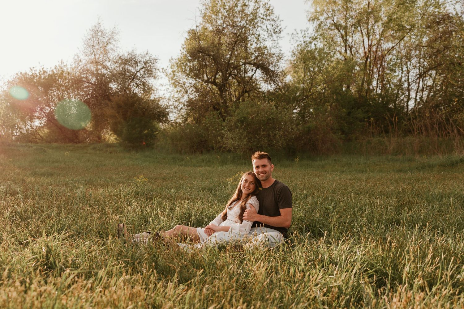Couple sitting together in a sunlit green meadow surrounded by trees during golden hour.