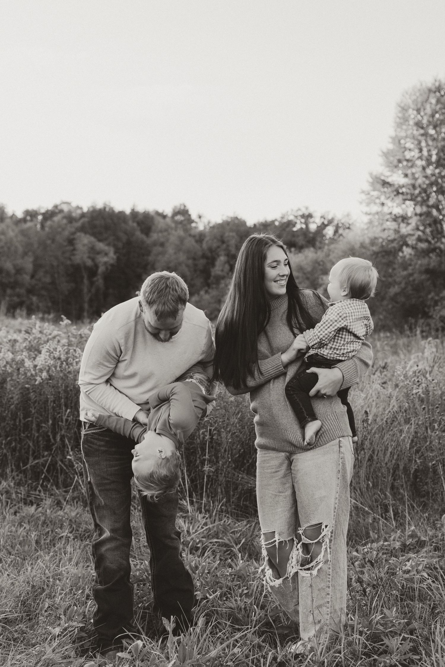 Black and white family photo of parents and baby in a field, mom holding infant while dad plays with toddler outdoors.