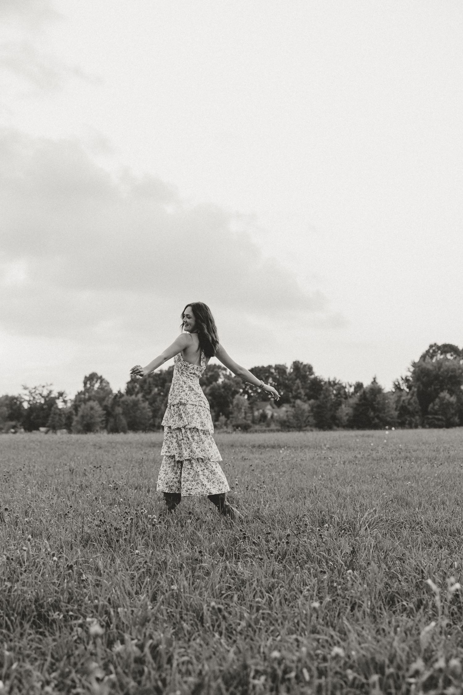 Woman in floral tiered maxi dress dancing freely in open green field, black and white photography.