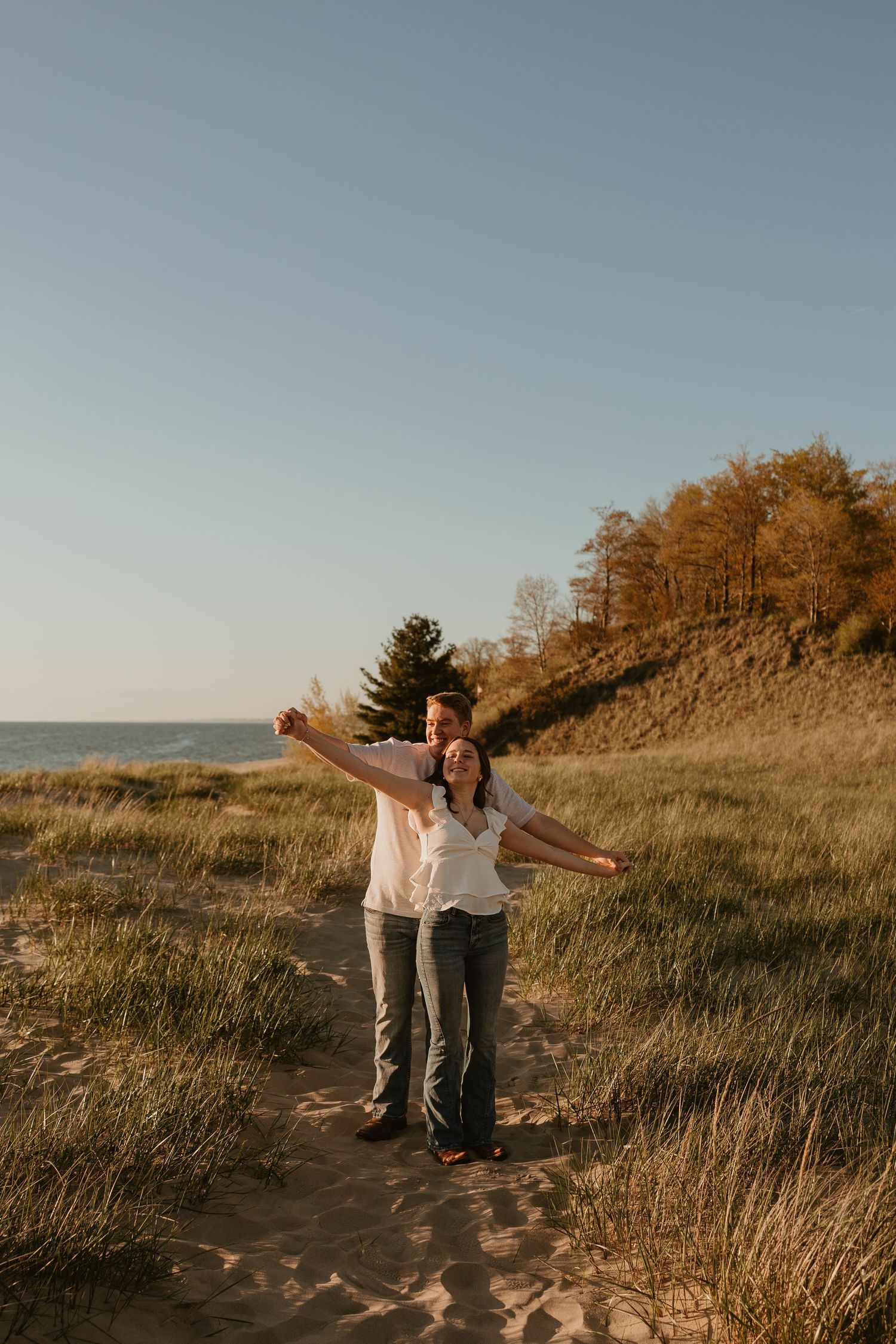 Couple joyfully posing on sandy beach path with arms outstretched, surrounded by golden autumn trees and ocean views.