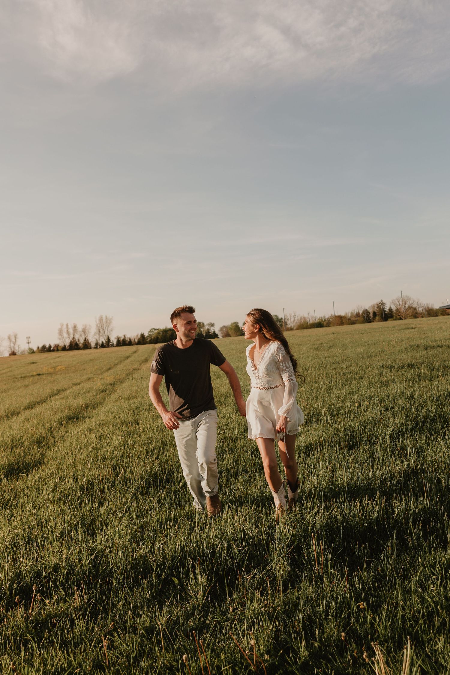 A couple walks hand-in-hand through a lush green field at sunset, smiling at each other.