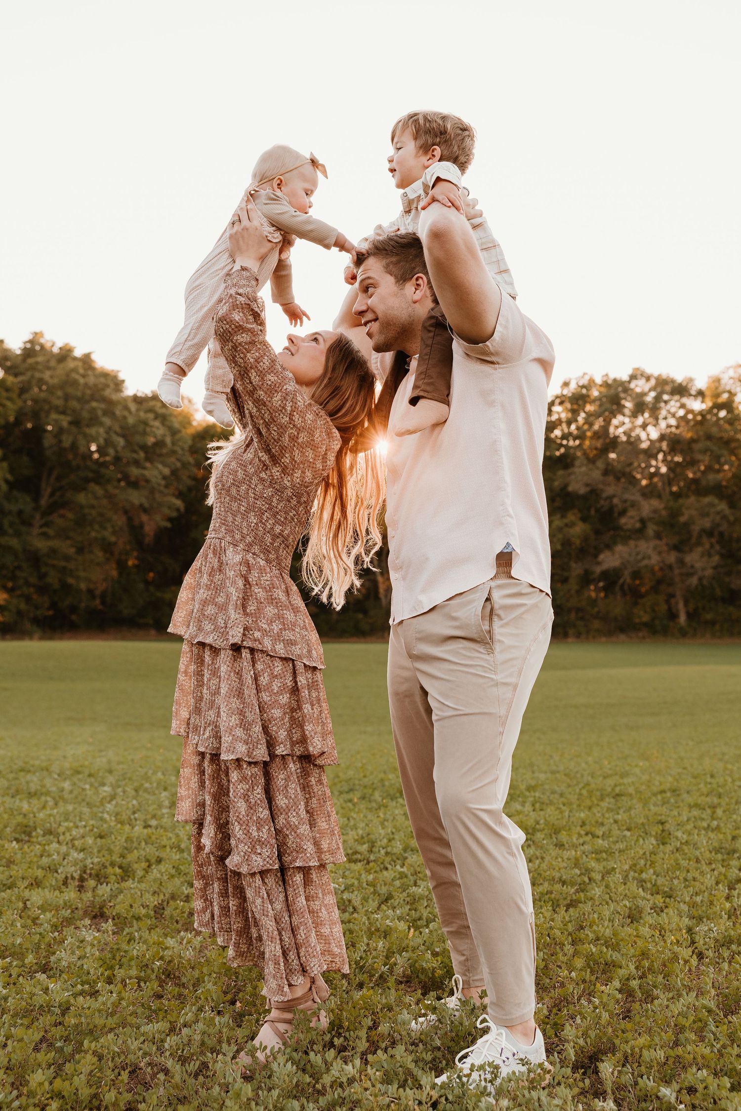 A joyful family of four plays outdoors at sunset, with parents lifting two young children in a lush green park.