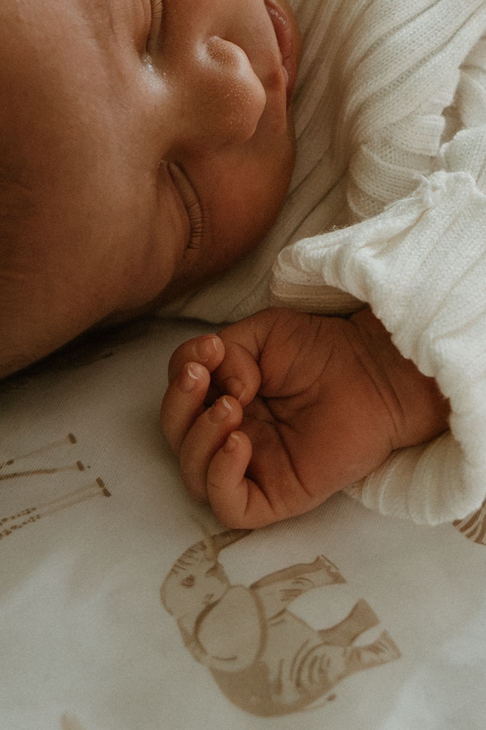 Sleeping newborn baby with tiny clenched fist resting on a white blanket with delicate animal print.
