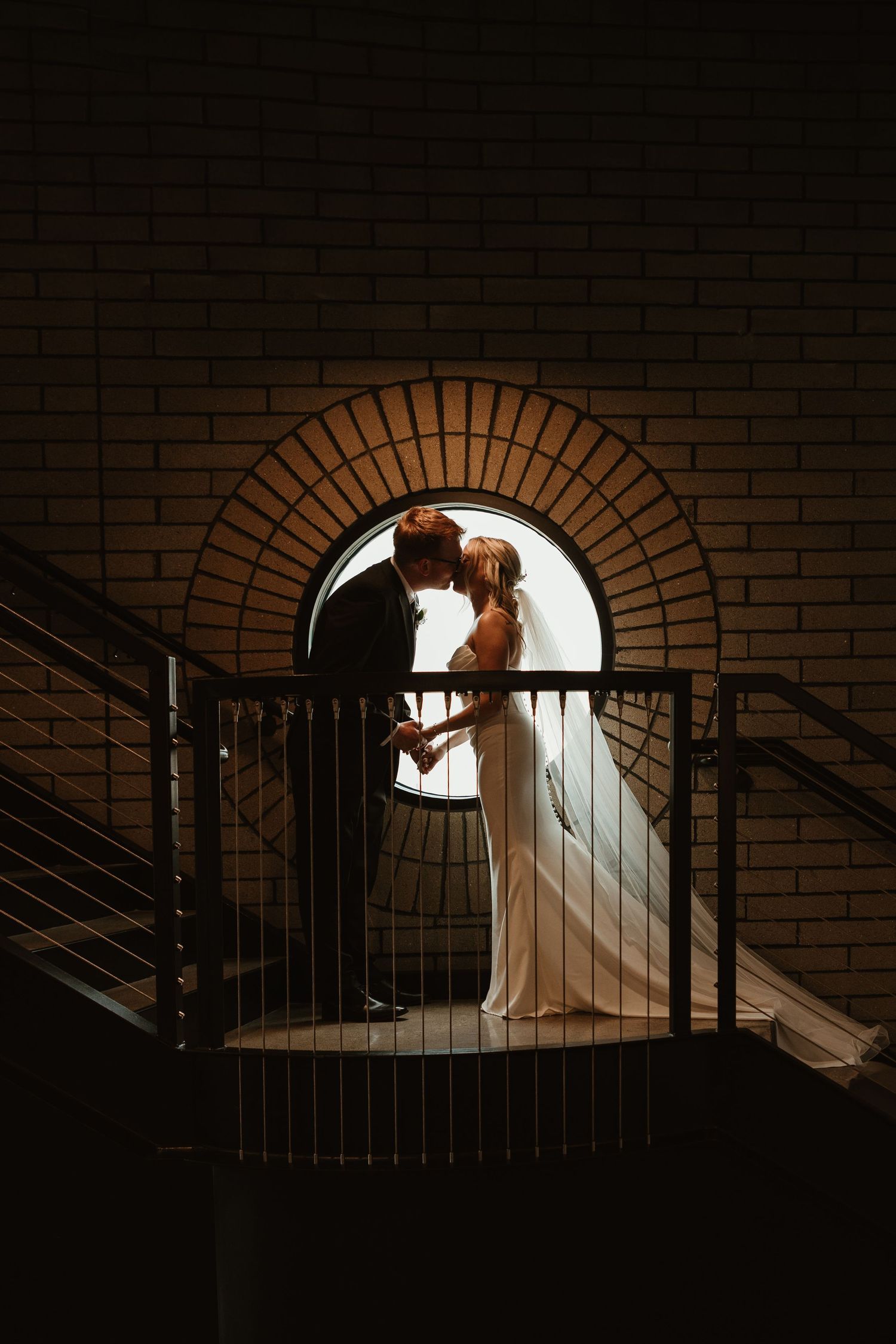 Bride and groom silhouetted in arched window on staircase landing, sharing romantic moment in dramatic dark setting.