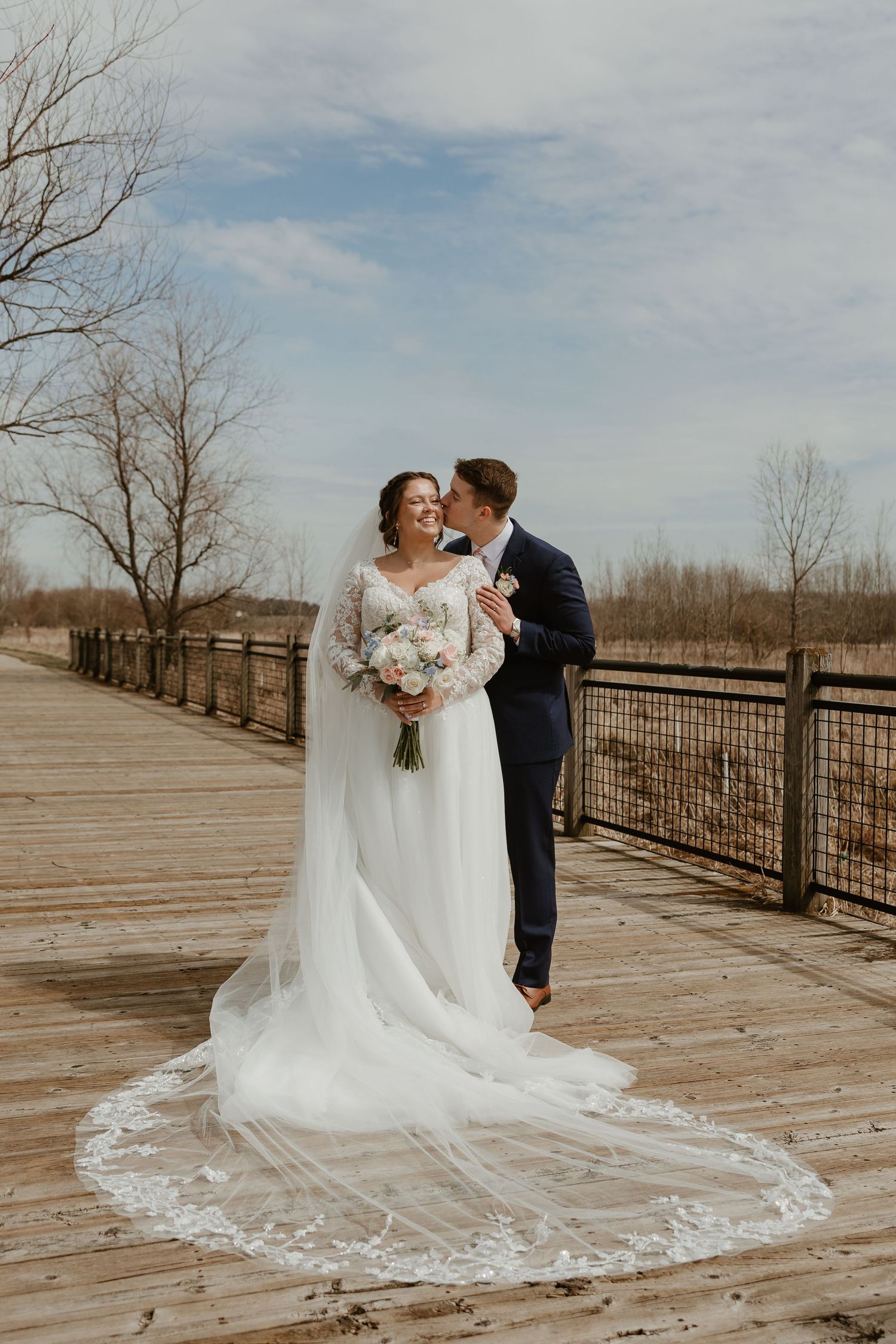 Bride in lace wedding gown with cathedral veil holds bouquet as groom kisses her cheek on wooden bridge.