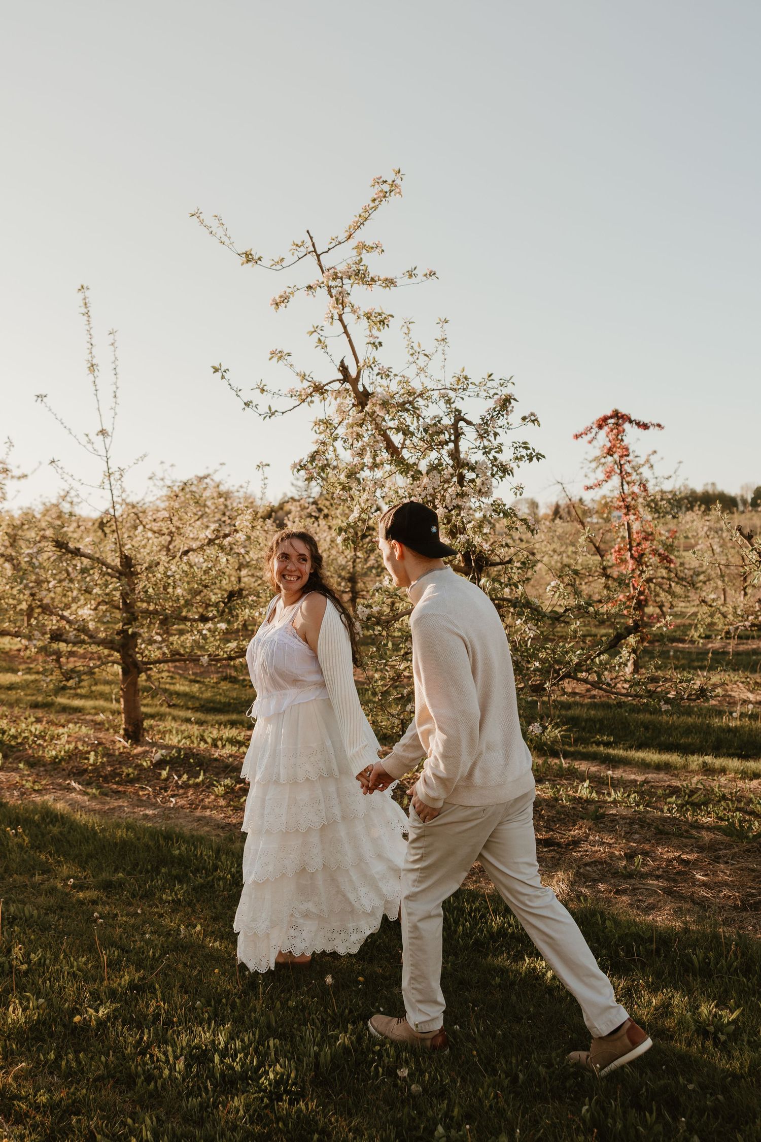 A bride in a white lace dress and groom in beige walk hand-in-hand through a blossoming orchard at golden hour.