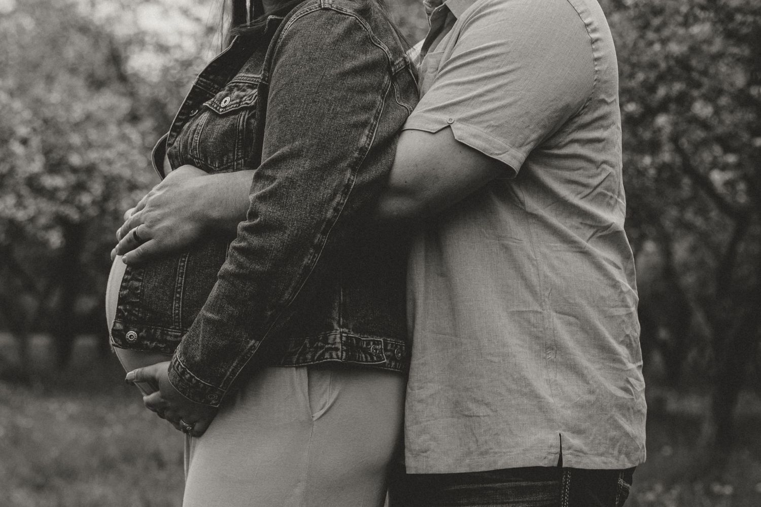 Black and white maternity photo of a couple embracing outdoors, showing pregnant belly in a garden setting.