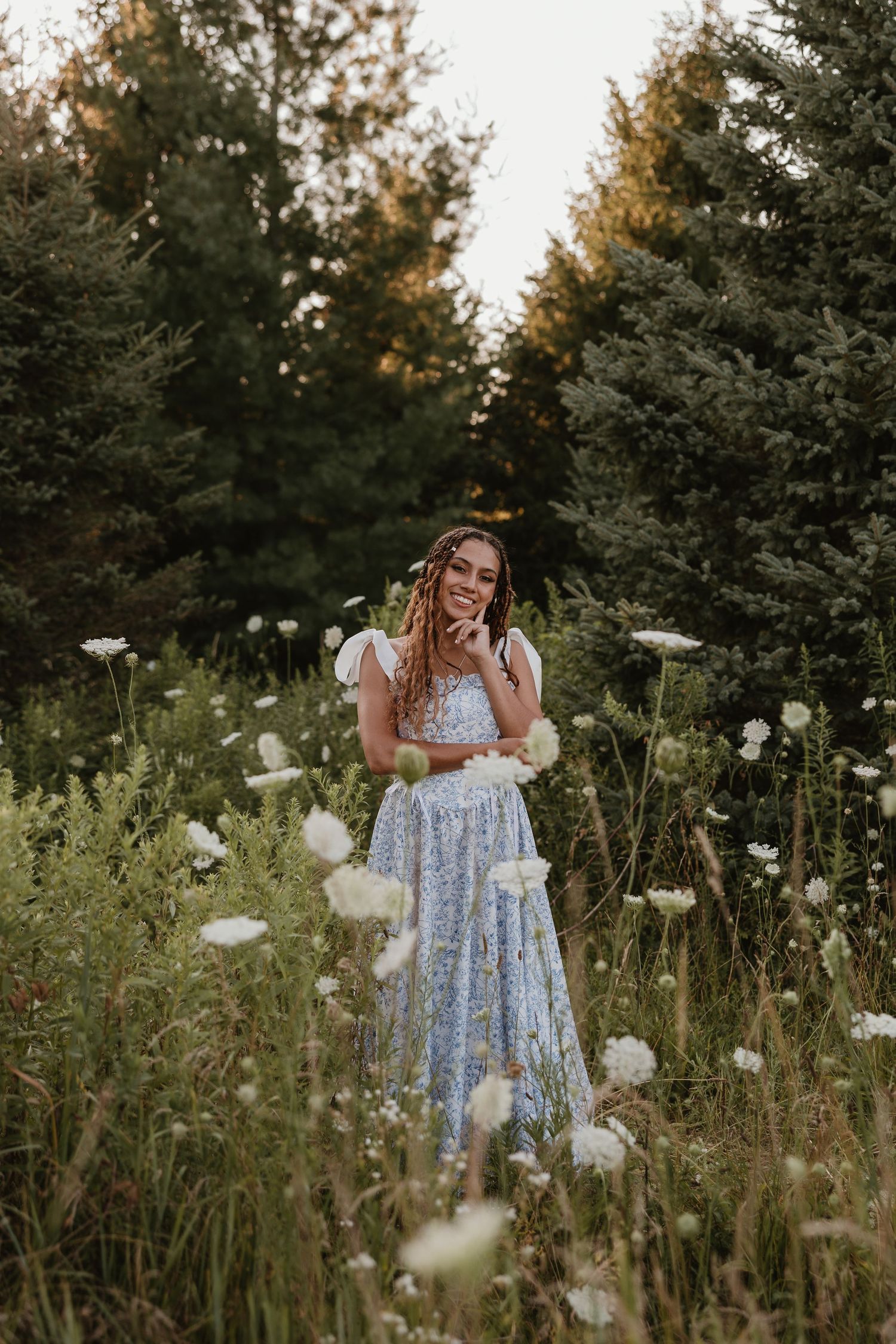 A smiling young woman in a blue floral dress stands among white wildflowers with evergreen trees in the background.