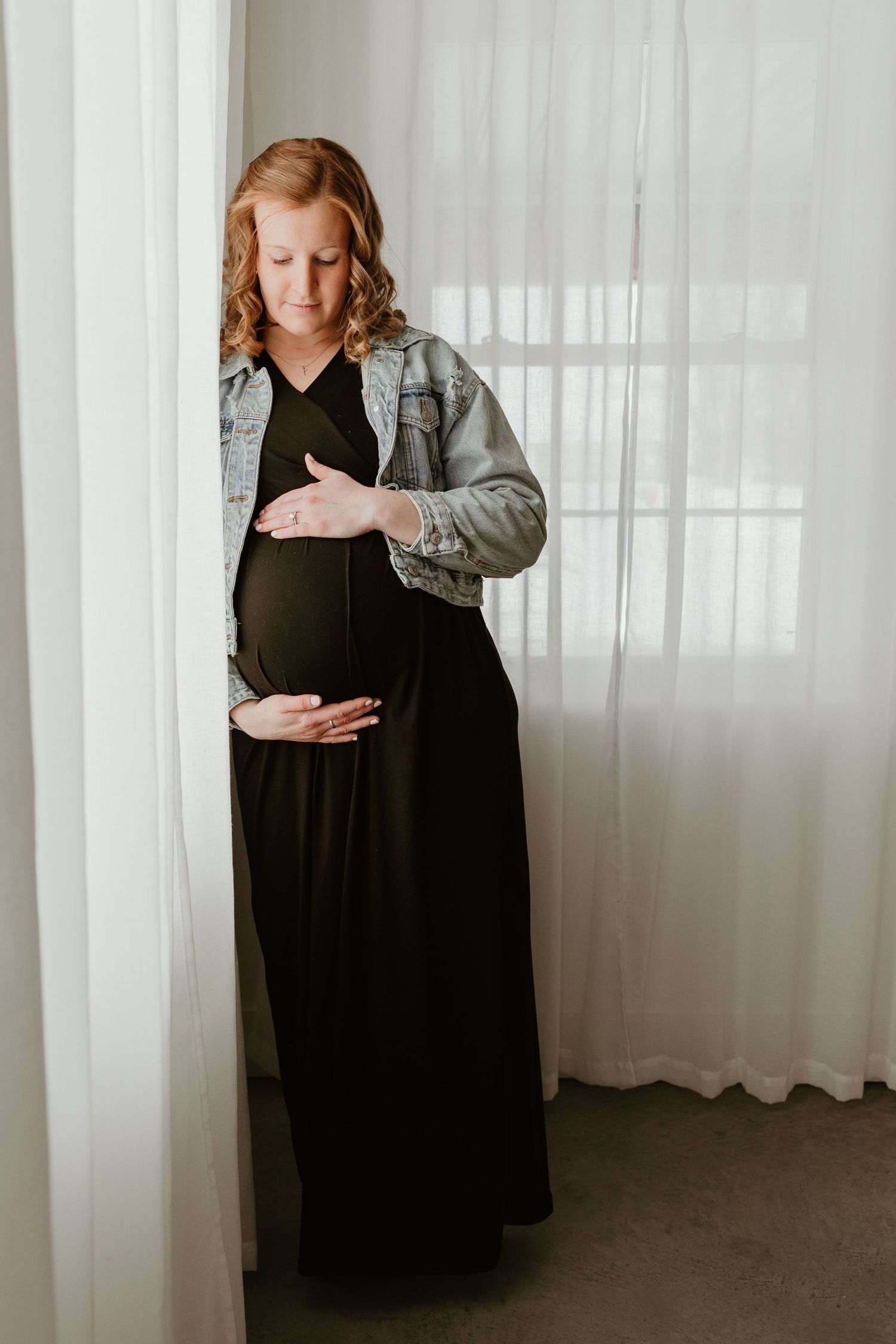 Pregnant woman in black maxi dress and denim jacket cradling baby bump by sheer white curtains.