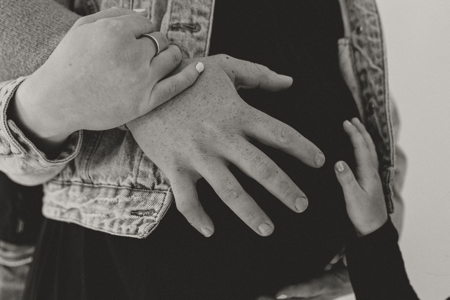 Black and white photo of two people embracing, toddler hands resting on a pregnant belly, during a family maternity session.