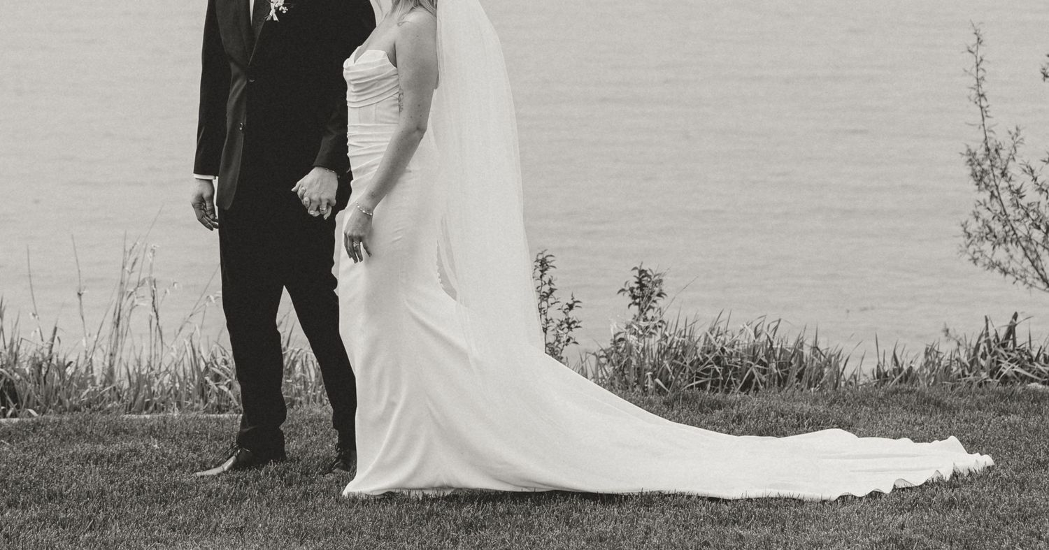 Black and white photo of a bride and groom holding hands by a lake on their wedding day.