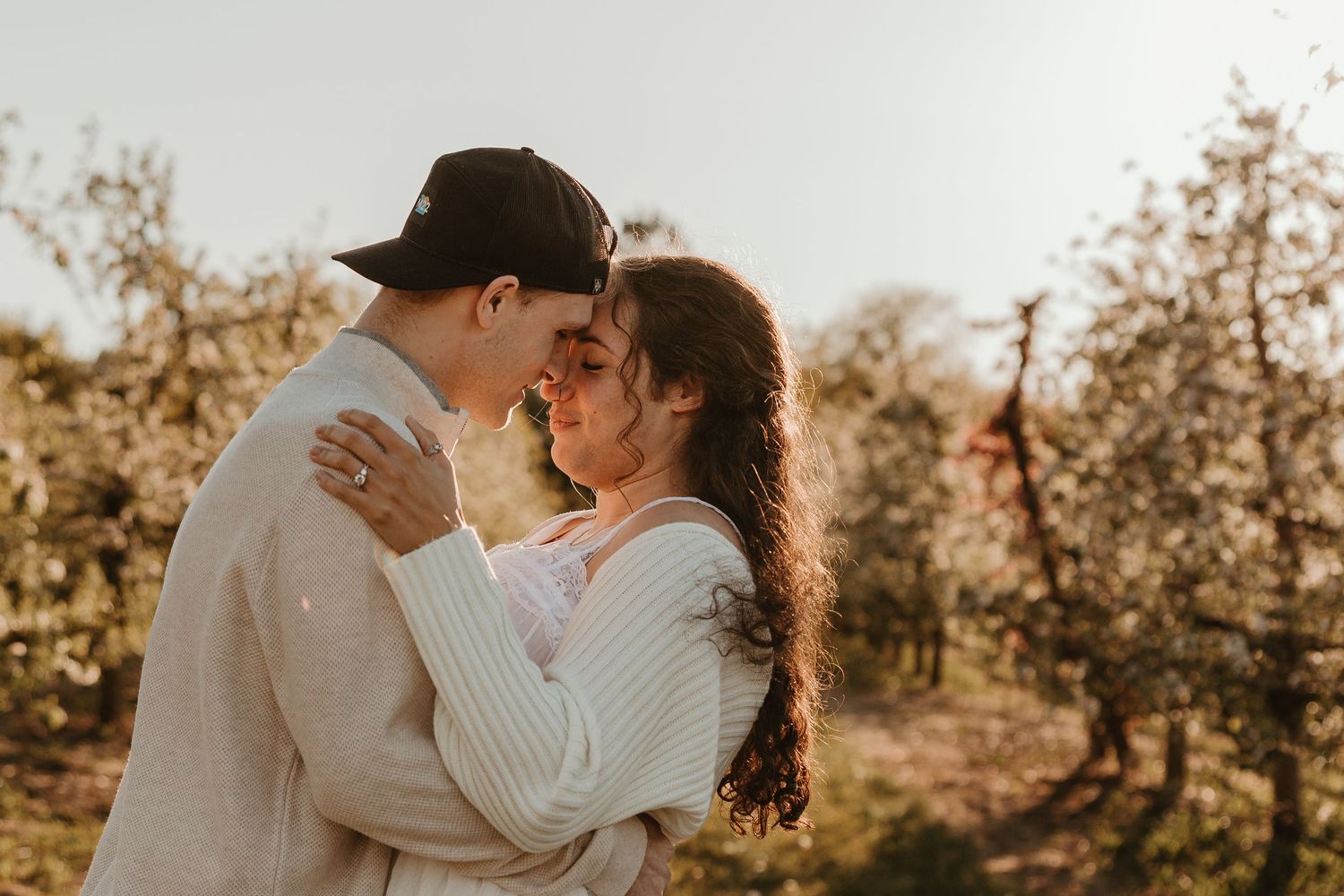 Romantic couple embracing and touching foreheads in a blooming orchard during golden hour sunset.
