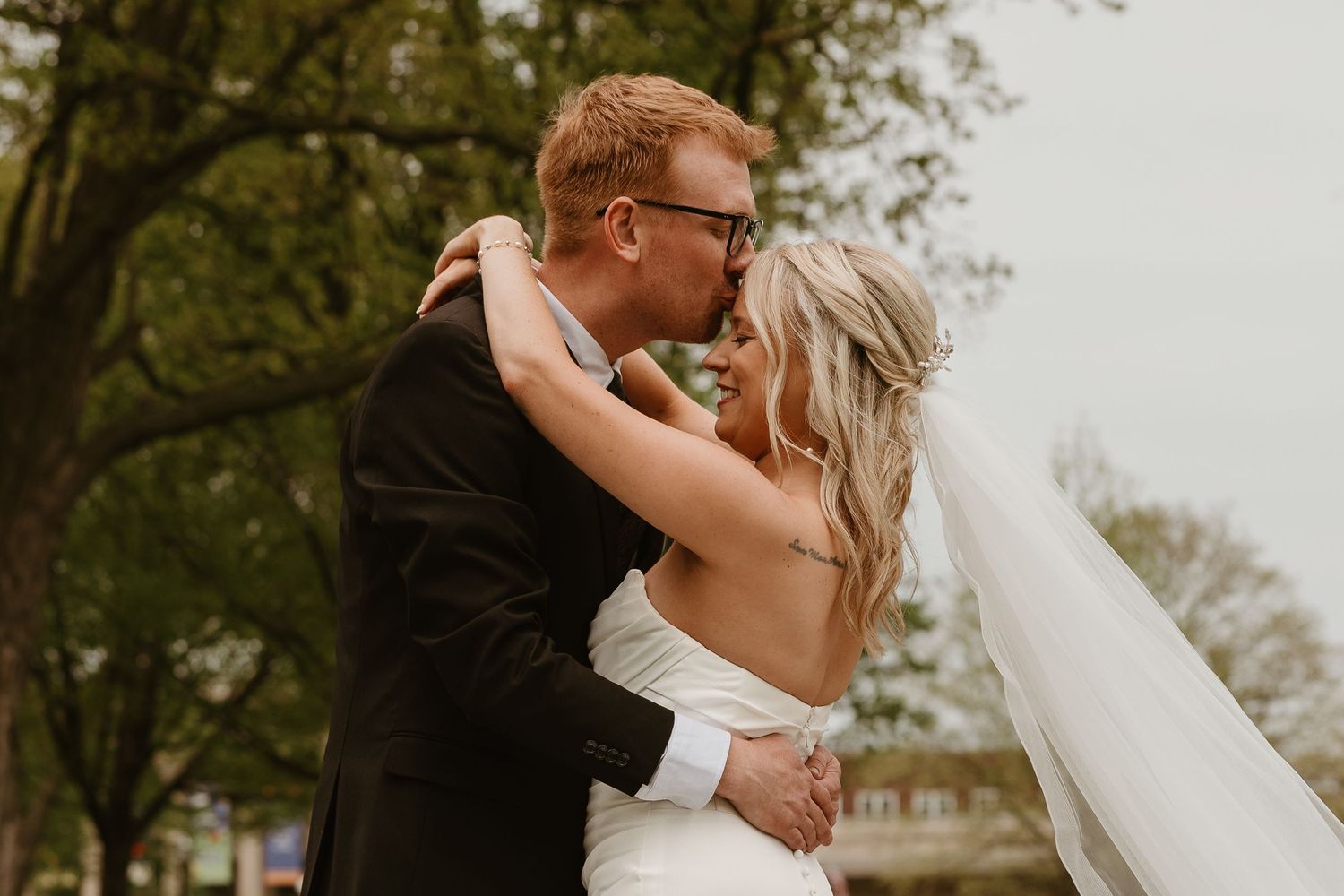 A bride and groom share a romantic kiss outdoors, with the bride's veil flowing in the breeze on their wedding day.