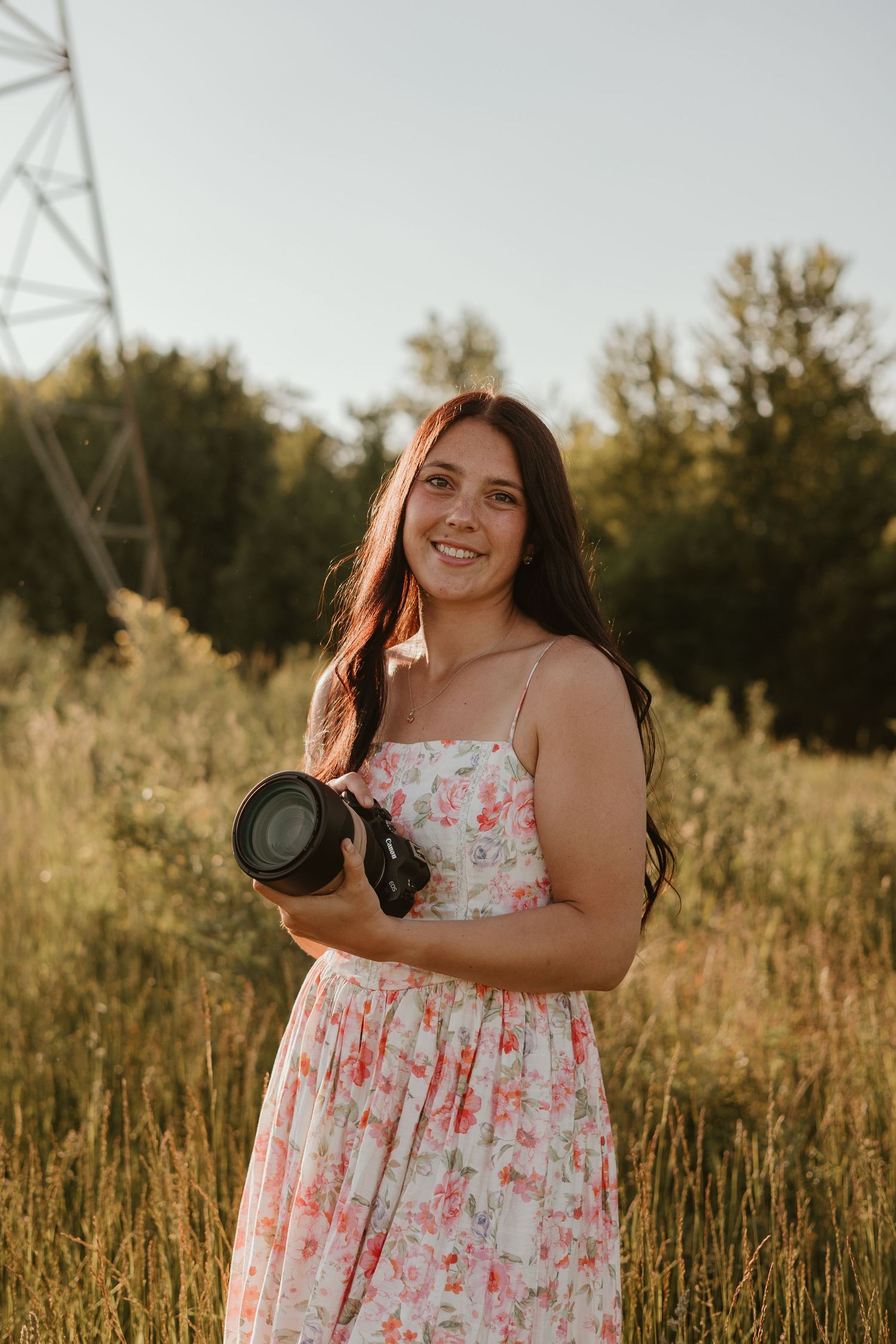 Young woman in floral dress holding a DSLR camera, smiling in a sunlit field with trees in the background.