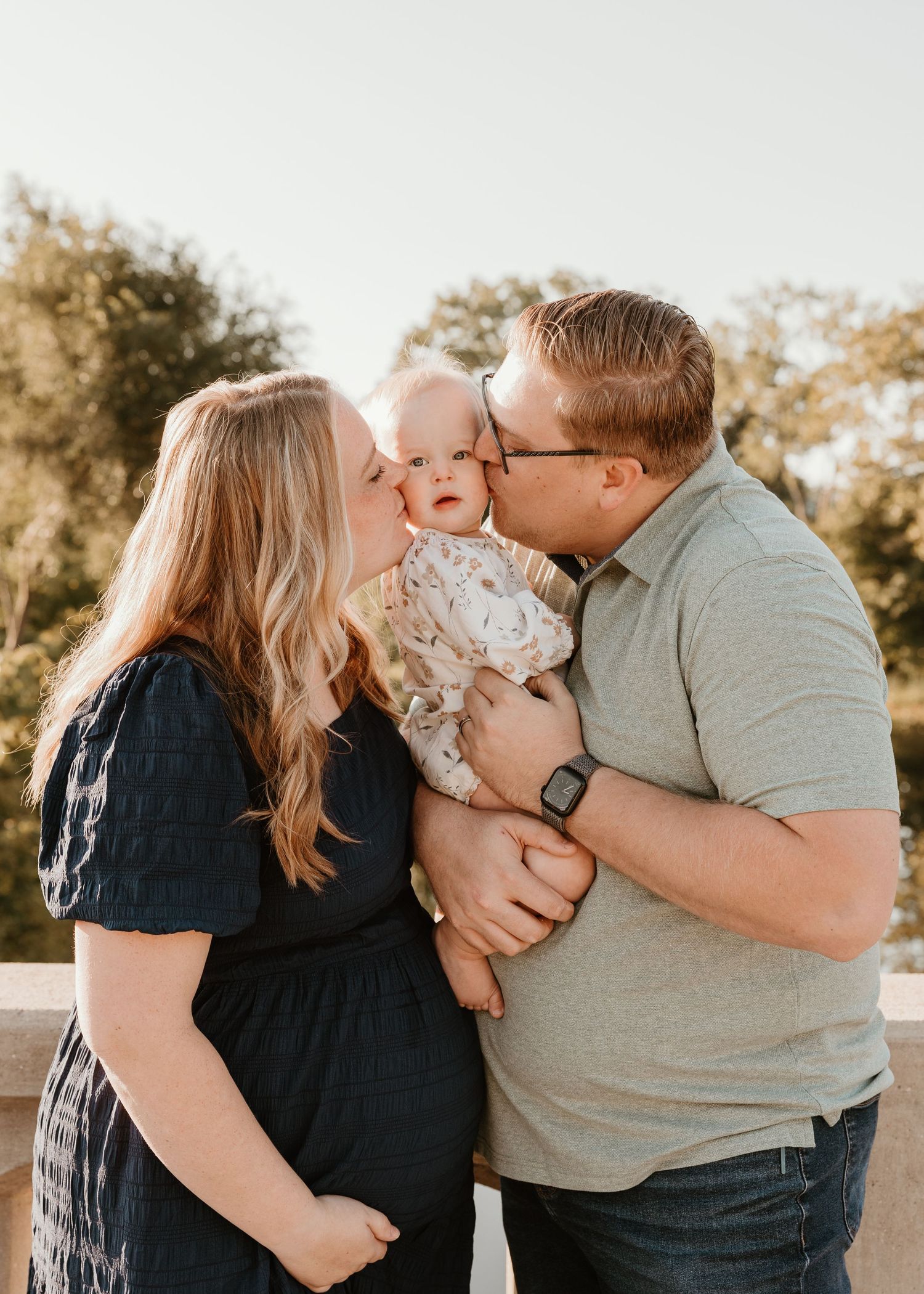 A couple kisses their baby on the cheeks during a golden hour outdoor family portrait session.