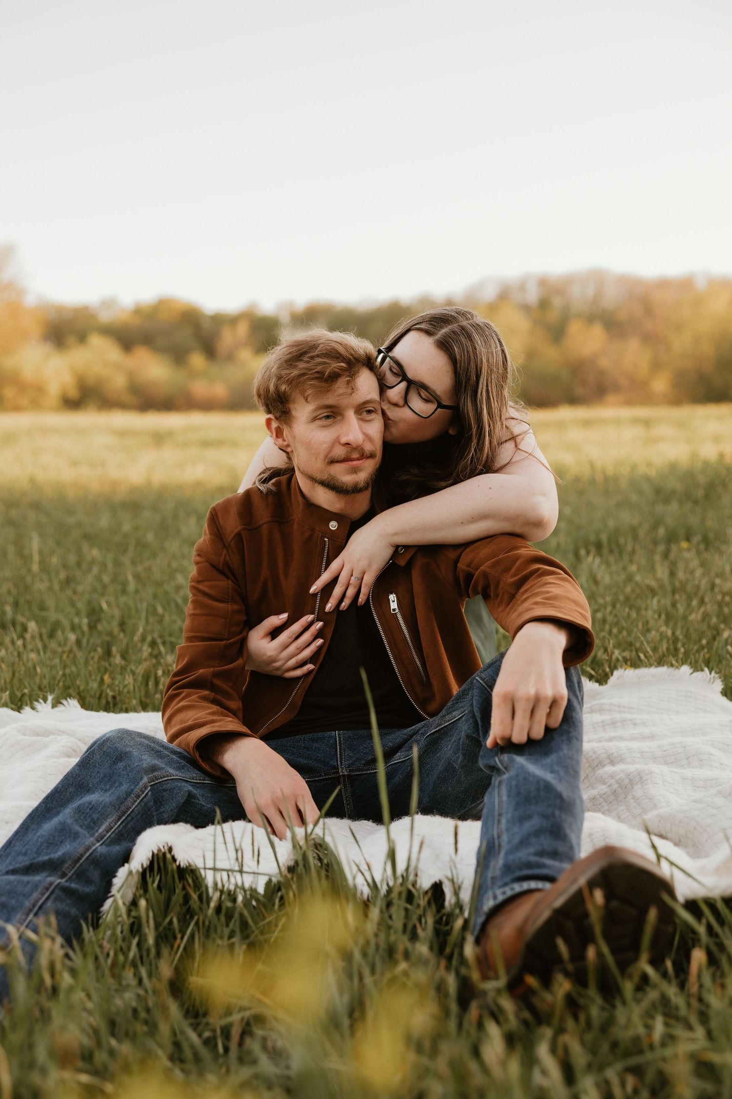 Couple sitting on a blanket in a green field, woman hugging man from behind, wearing casual fall outfits outdoors.