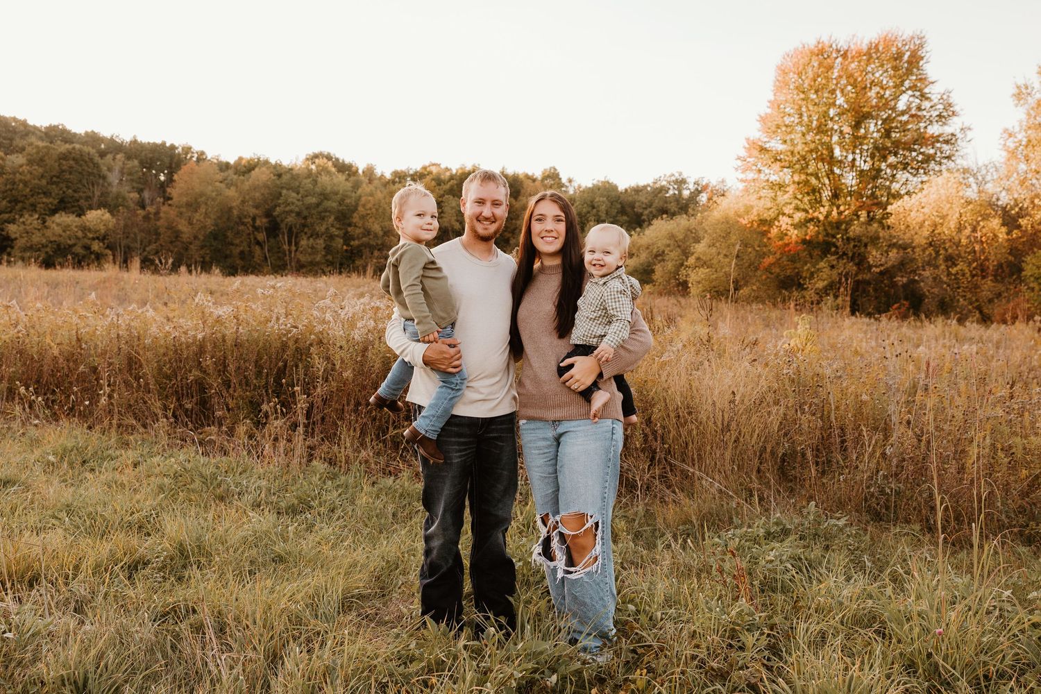 Family of four posing in a golden autumn field, parents holding two young children, smiling outdoors.