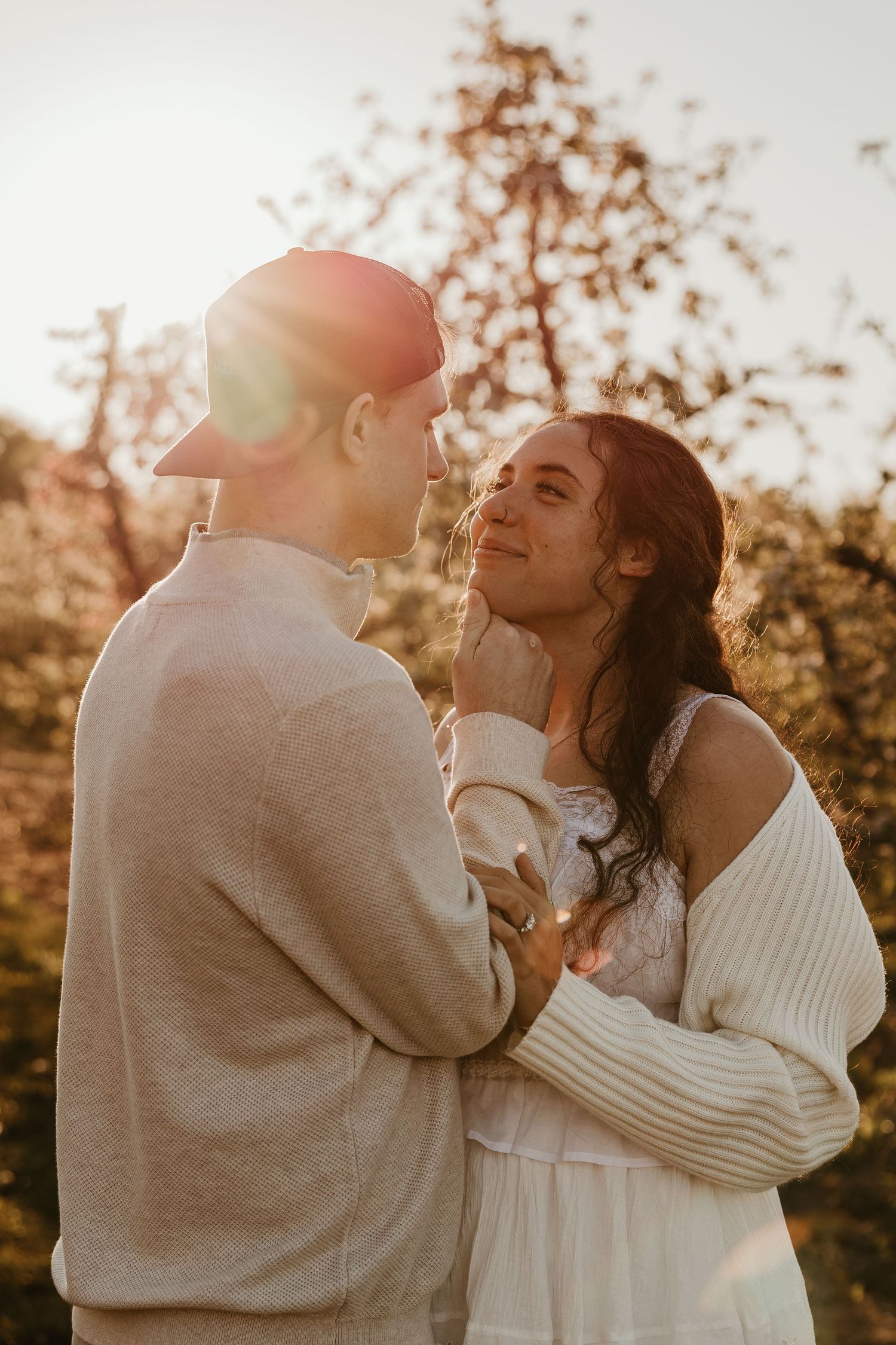 A couple shares a tender moment at golden hour, with warm sunlight and autumn trees in the background.
