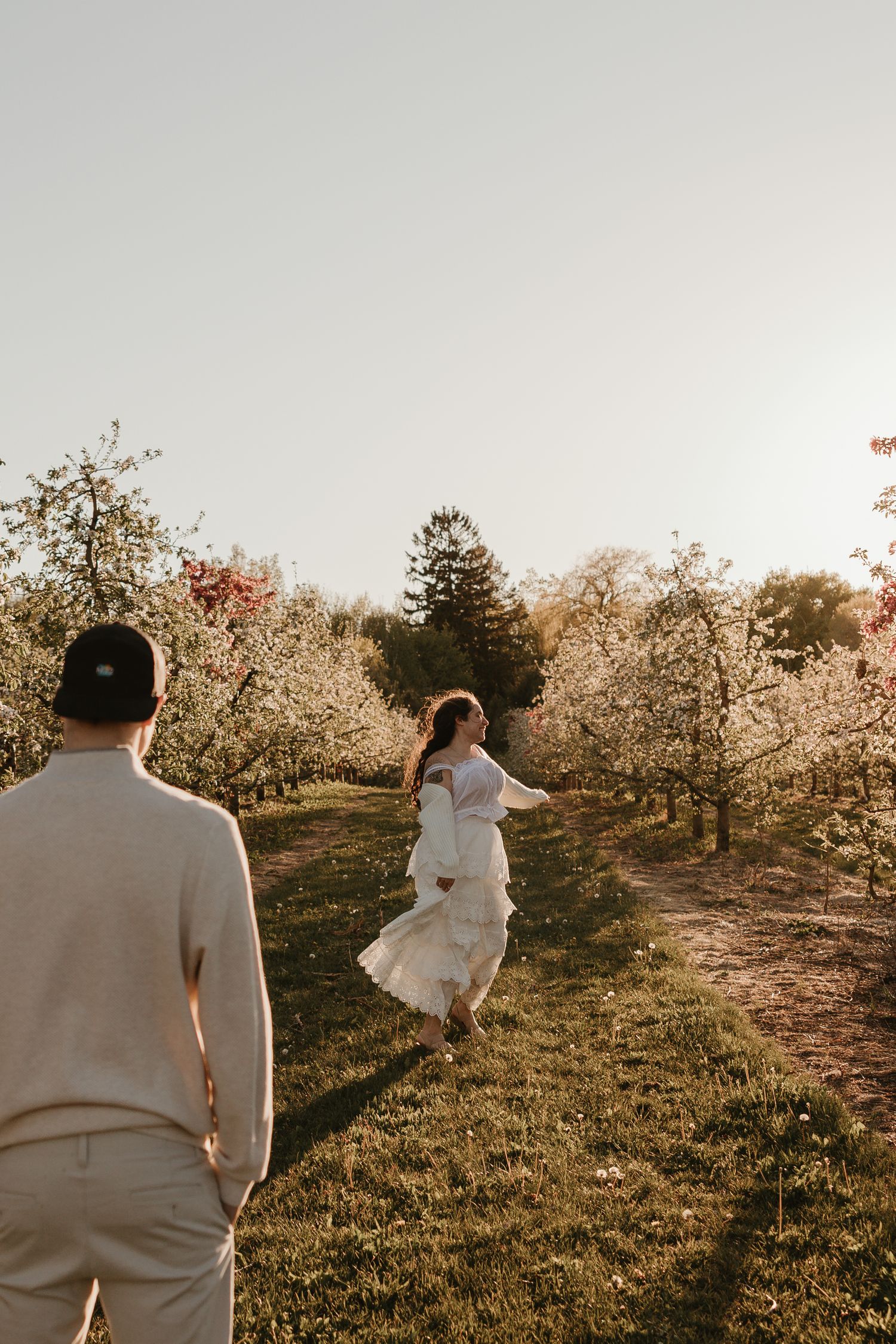 A bride in a white dress twirls in a sunlit orchard while her groom watches during a golden hour engagement session.