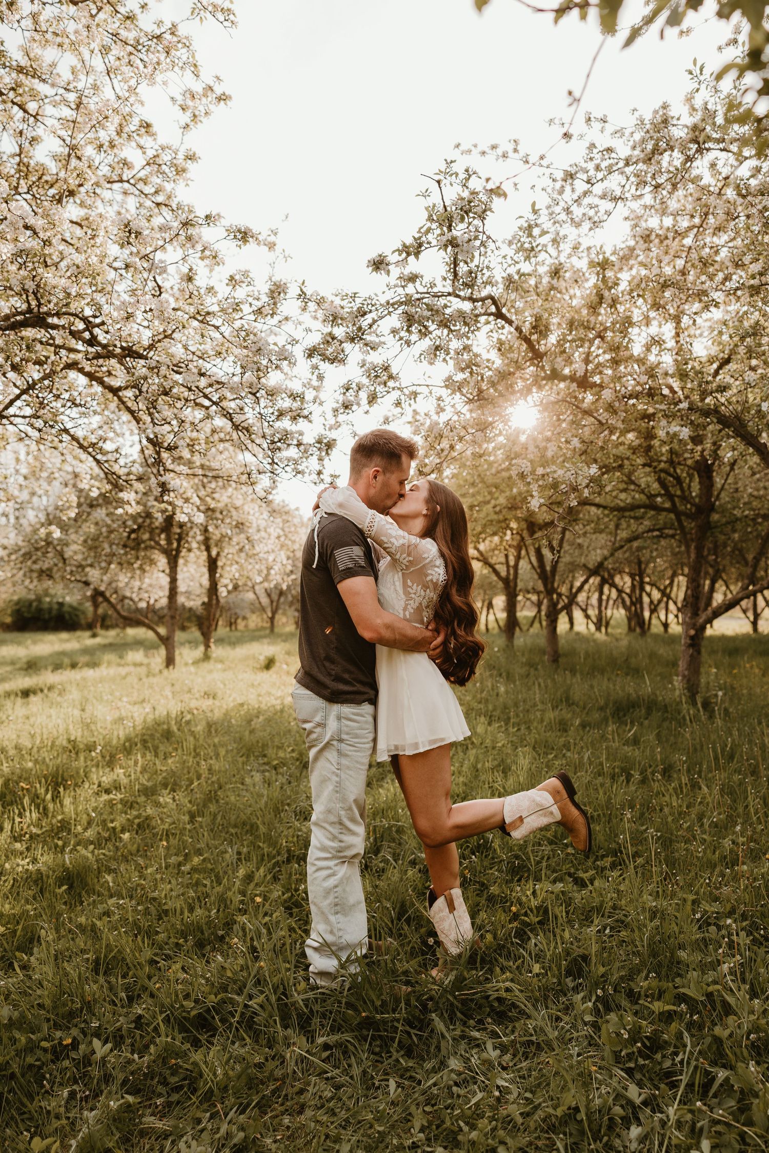 Romantic couple kissing in a blooming spring orchard at golden hour, woman in white dress lifting her leg.