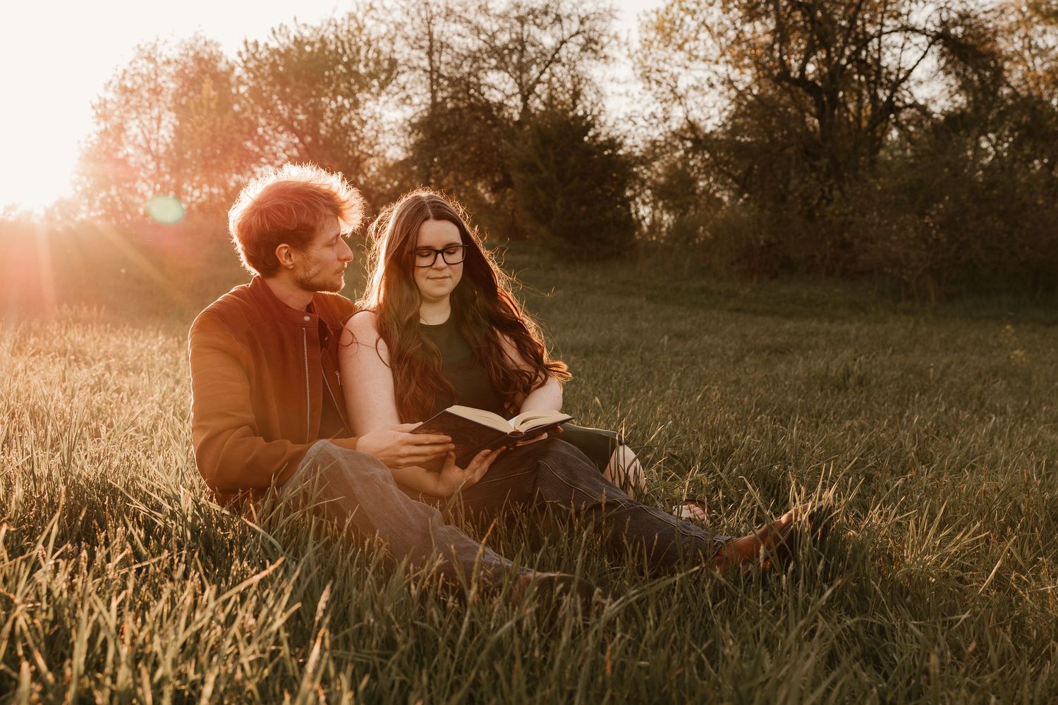 Couple sitting in golden hour sunlight in a meadow, woman reading a book while man looks on warmly.