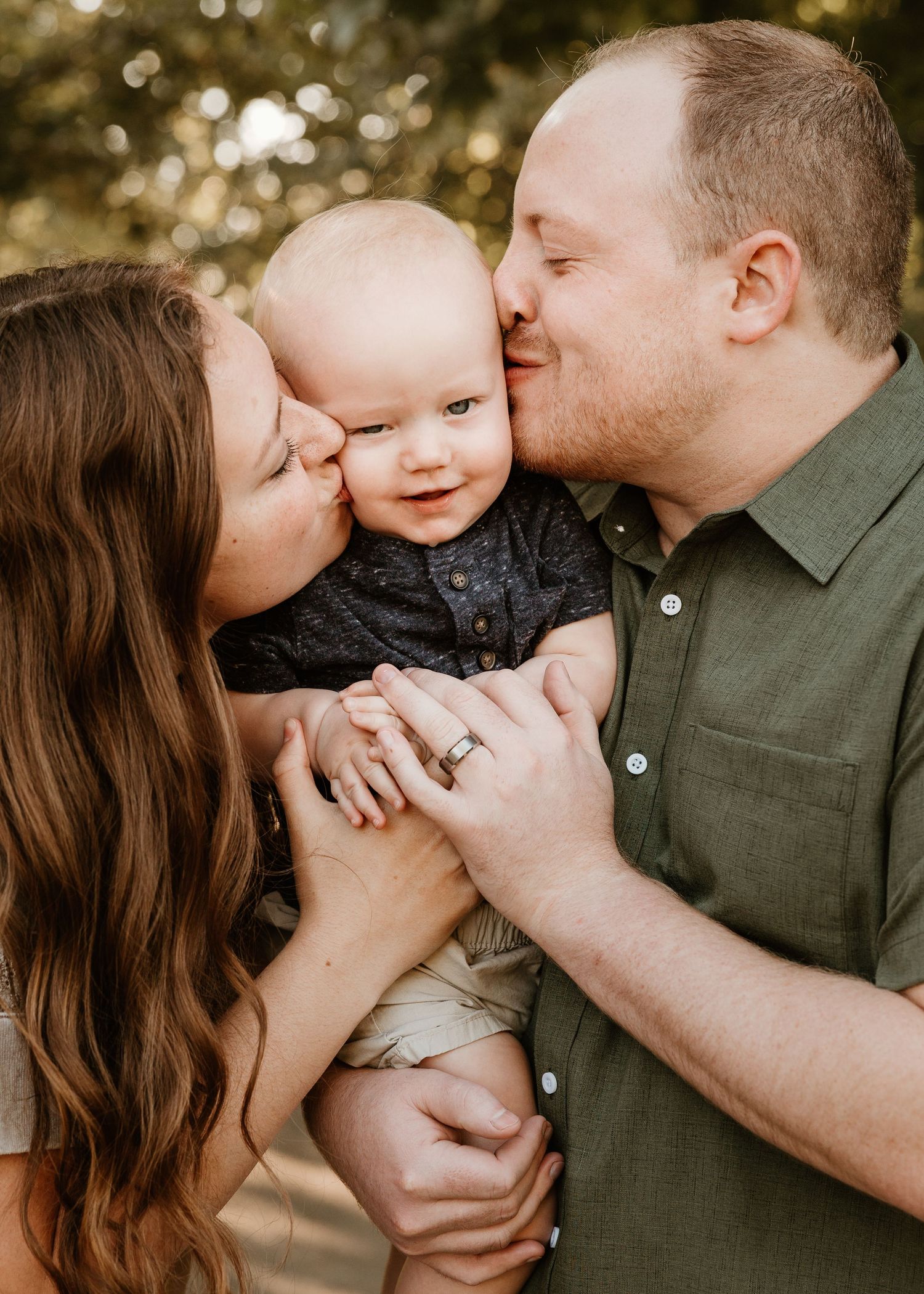 A young couple kisses their smiling baby on the cheeks during a warm outdoor family portrait session.