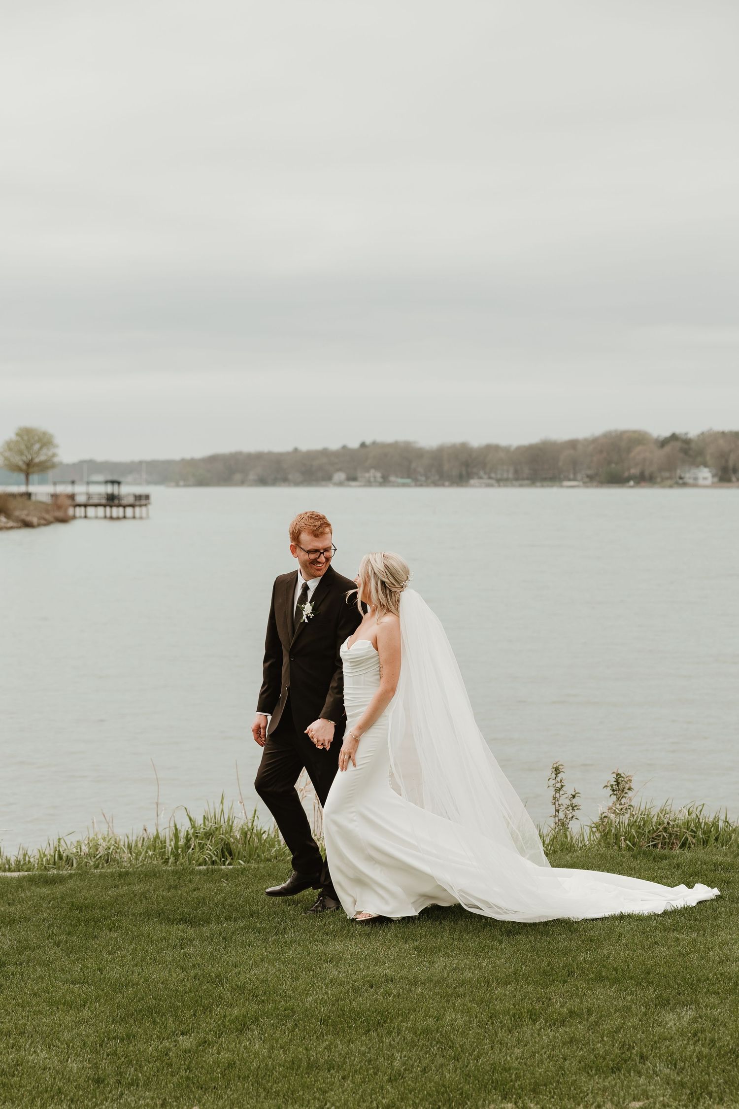 A bride and groom walk hand in hand on a green lawn beside a serene lake on their wedding day.