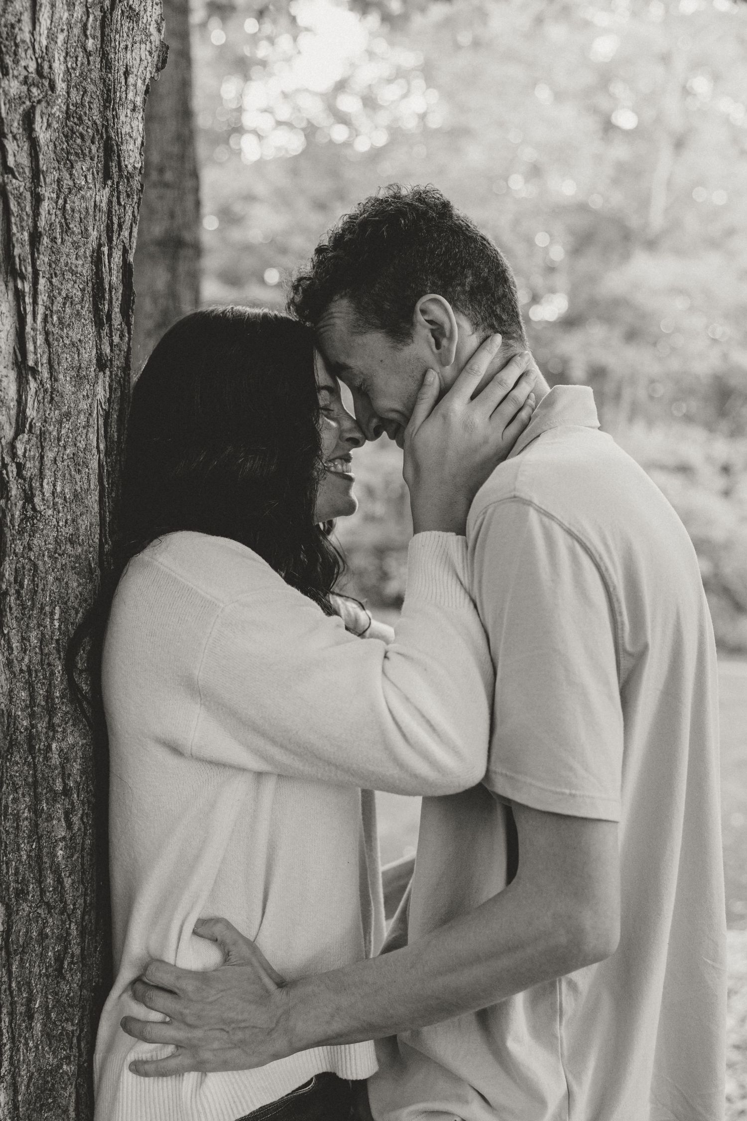 Black and white photo of a romantic couple embracing and almost kissing beside a tree in a park.