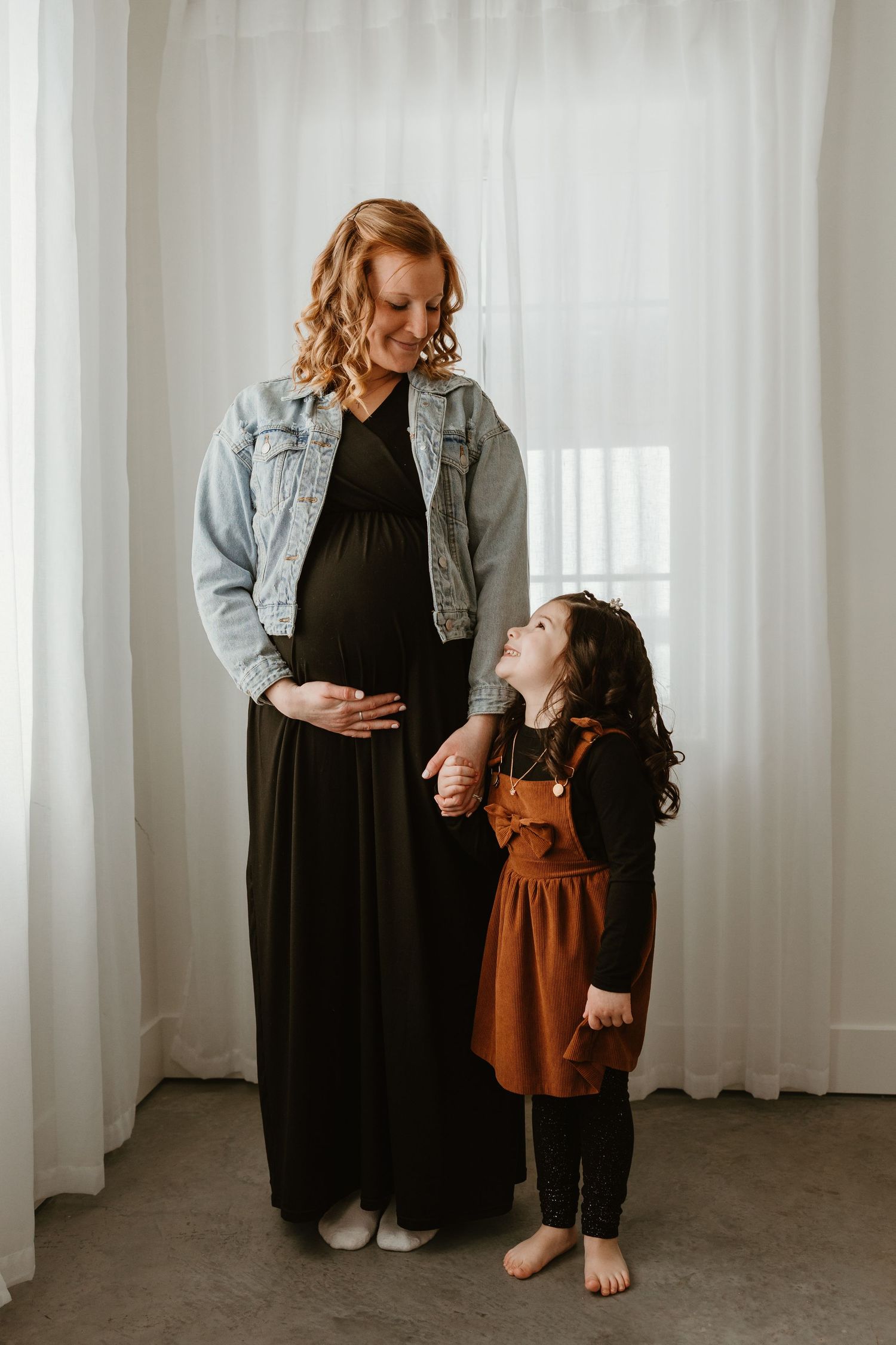 Pregnant woman in black dress and denim jacket holds hands with young girl in rust dress during maternity photo session.