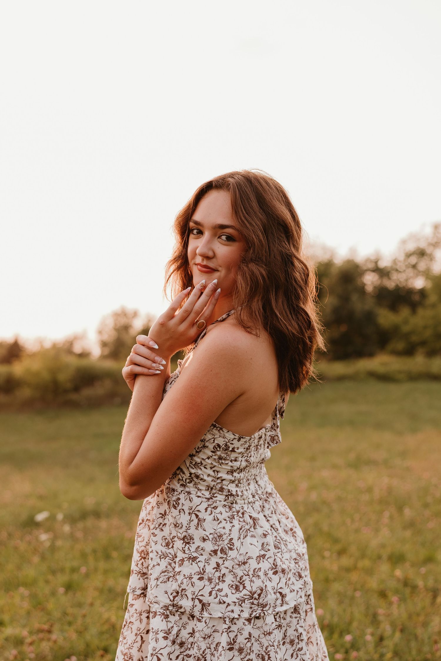 Young woman in floral sundress poses gracefully in a golden hour field, smiling over her shoulder.