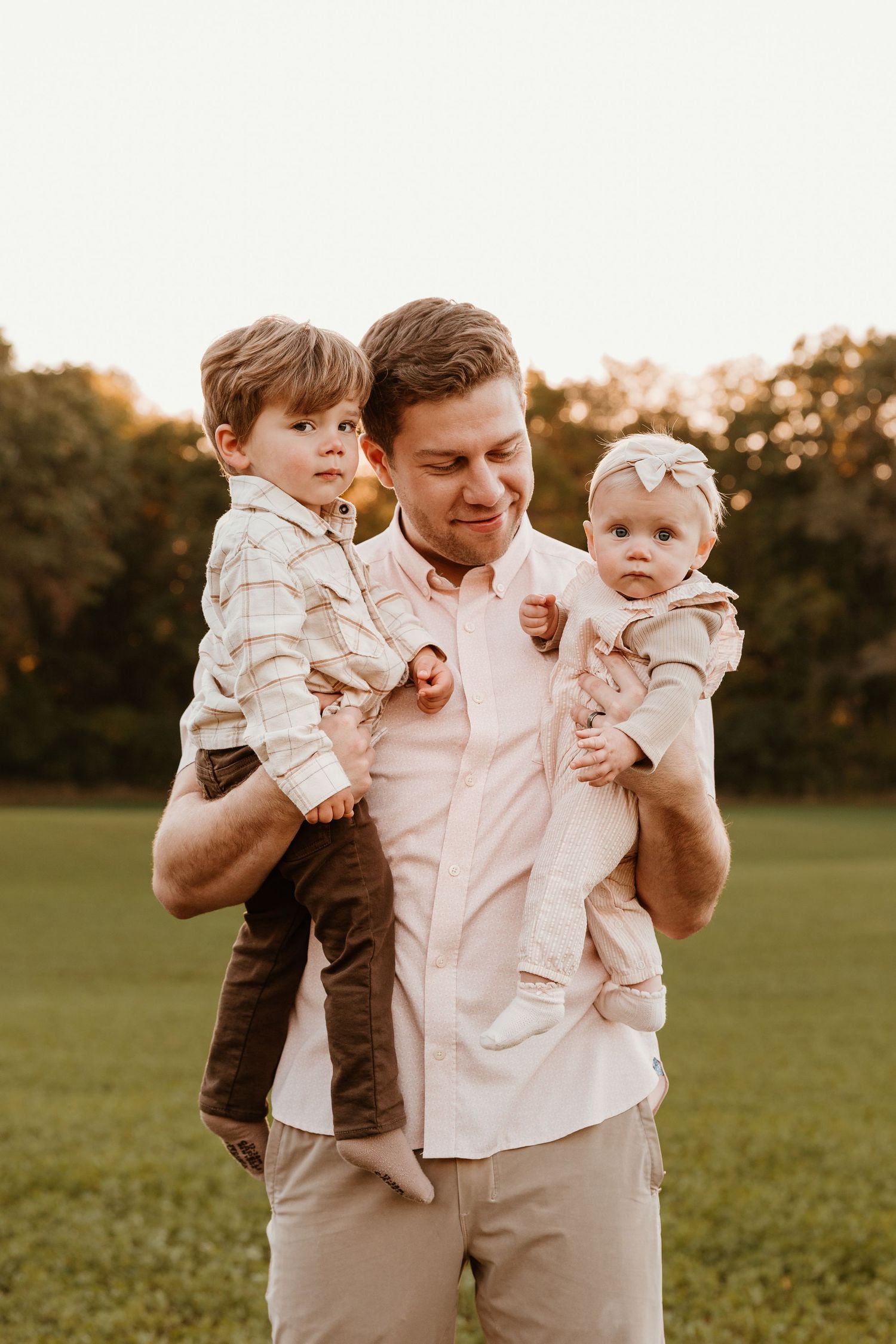 Father holding two young children in a sunny green field during golden hour family portrait outdoors.