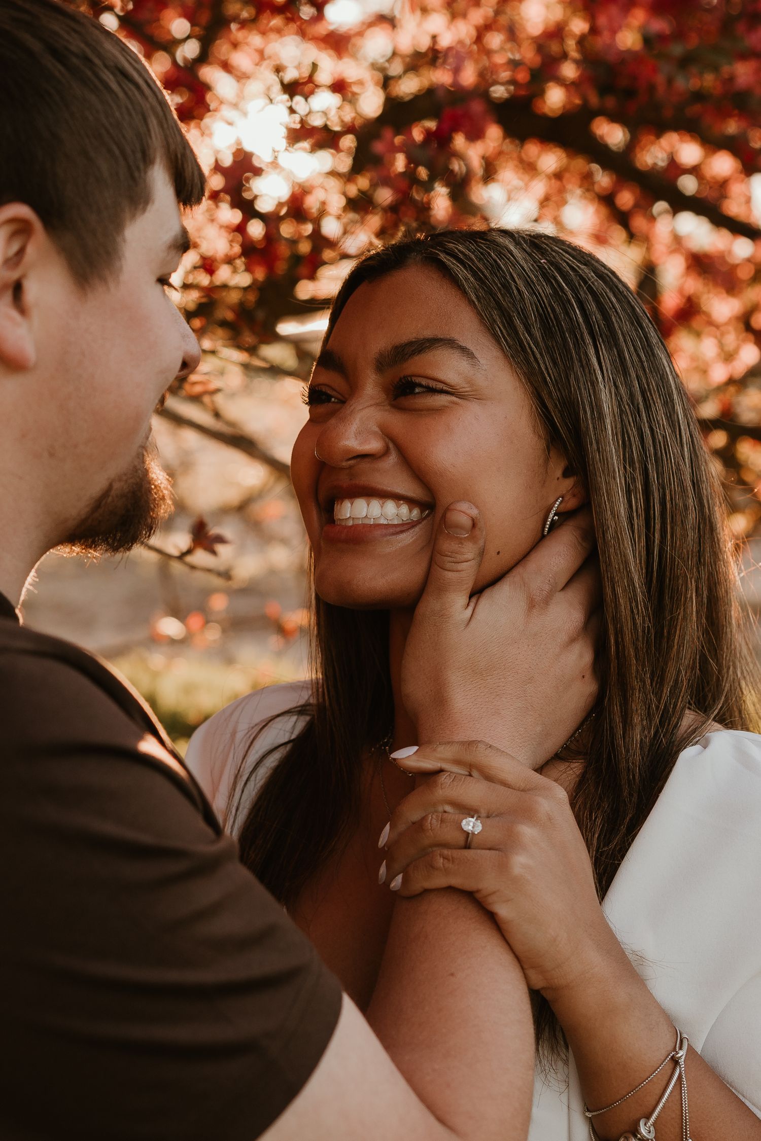 A joyful woman smiles as her partner gently holds her face, wearing an engagement ring, surrounded by autumn foliage.