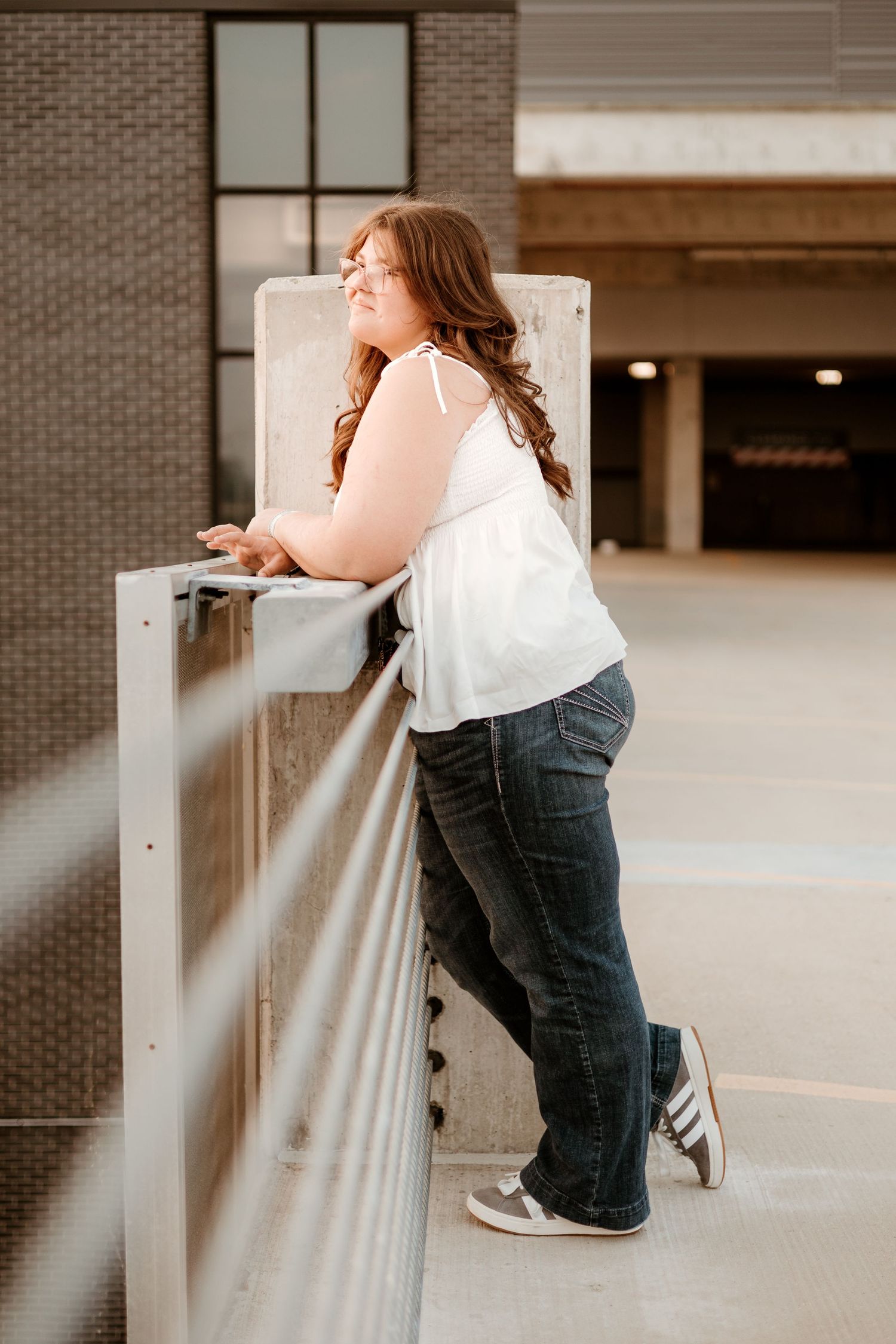 Young woman in white top and jeans leans on metal railing at parking garage, smiling sideways.