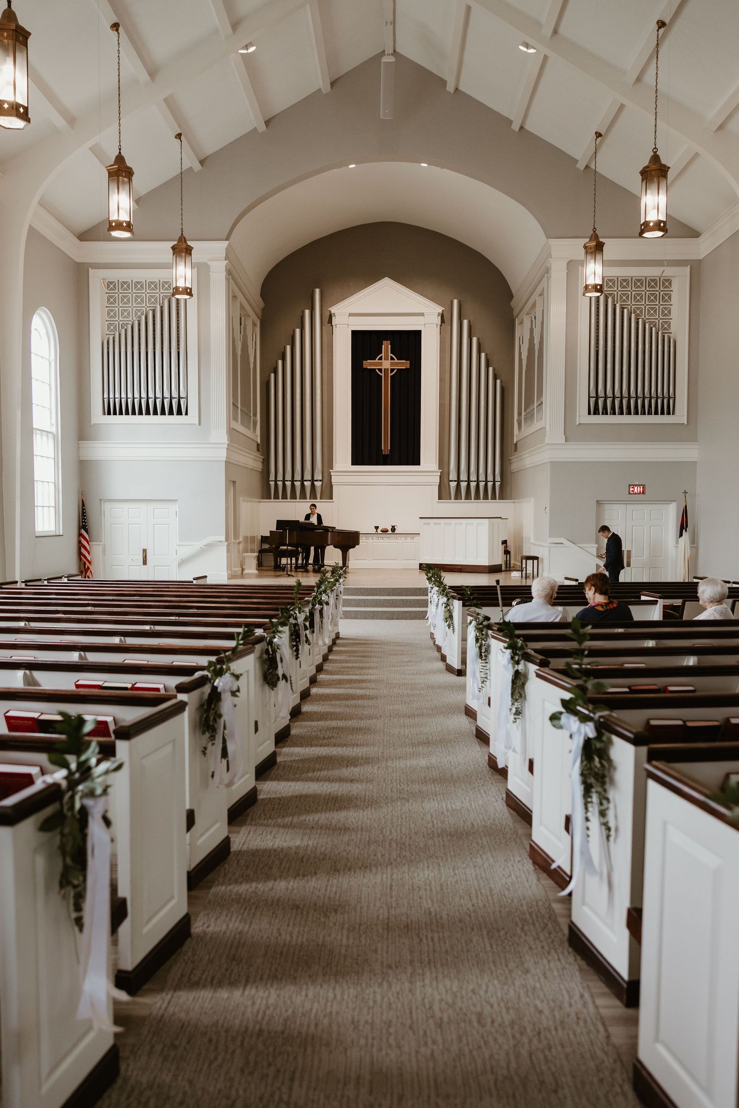 Elegant church interior with white pews adorned with greenery, pipe organs, and a wooden cross at the altar during a wedding.
