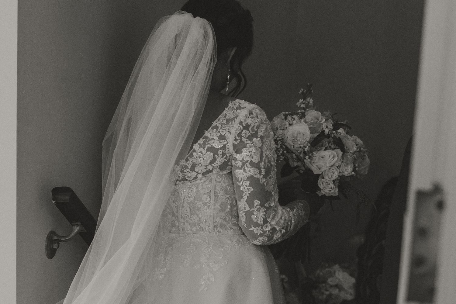 Black and white photo of a bride in lace gown and veil holding a bouquet, viewed from behind.