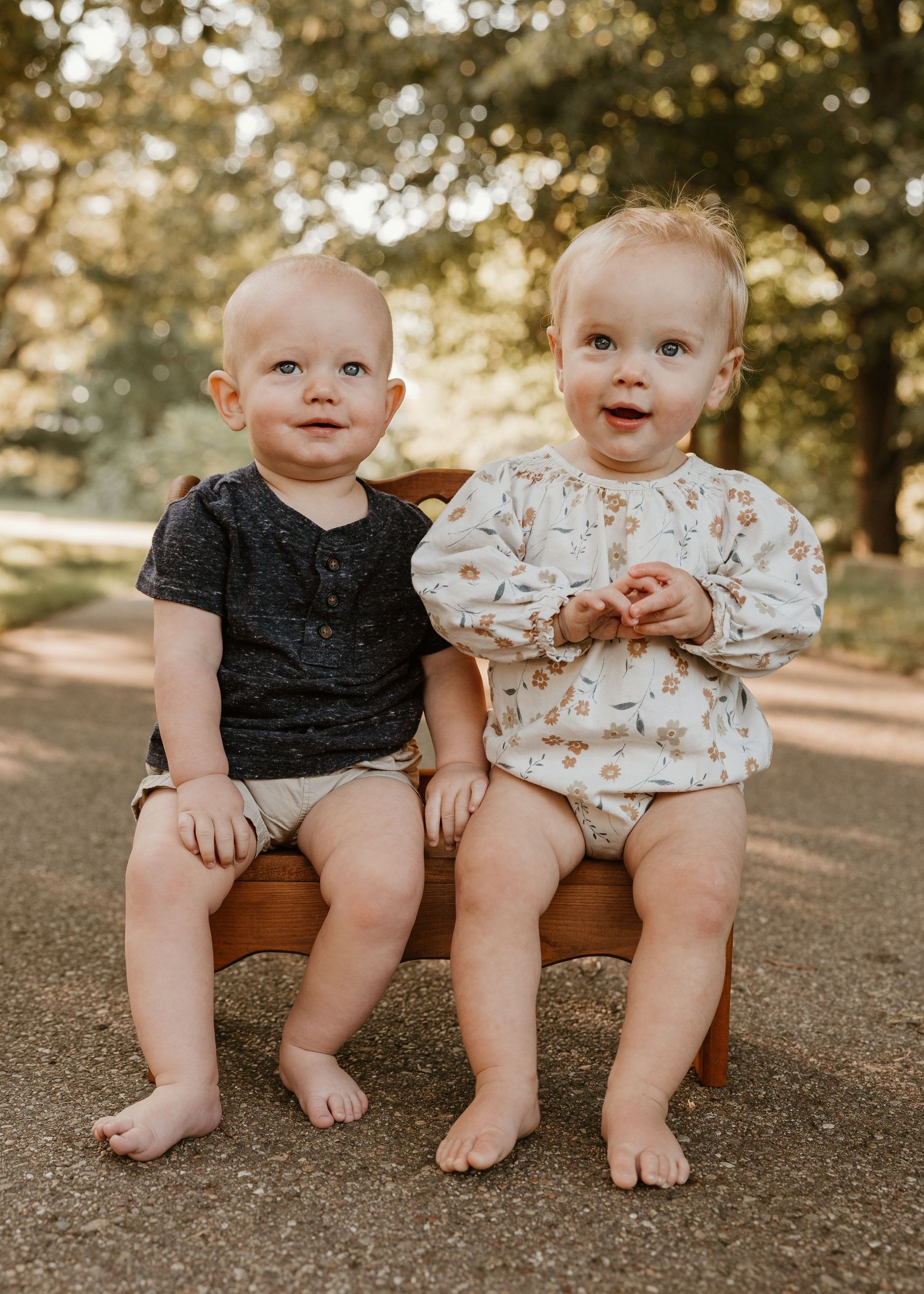 Two adorable babies sitting together on a small wooden bench outdoors, smiling in natural light.