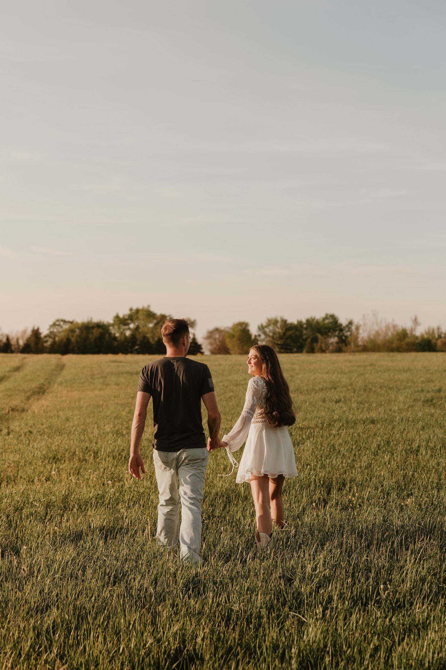 A couple holds hands while walking through a lush green field at golden hour, woman in white dress looks back smiling.