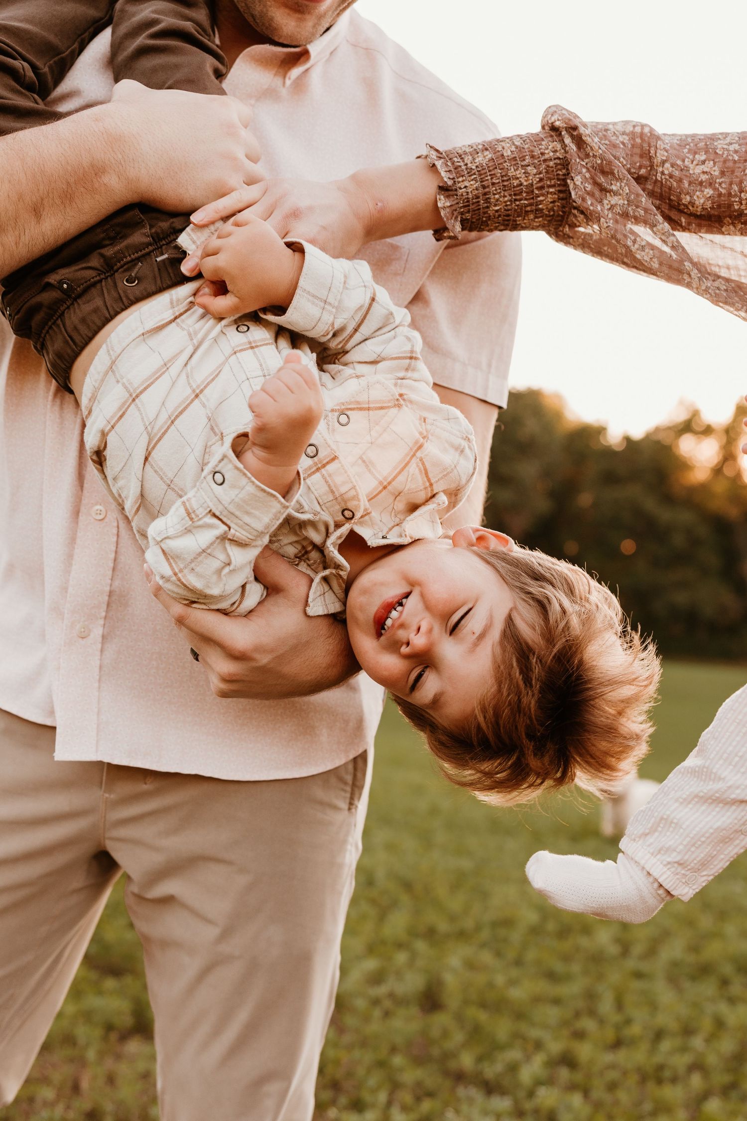 Parents playfully holding laughing toddler boy upside down in a sunny outdoor field at golden hour.