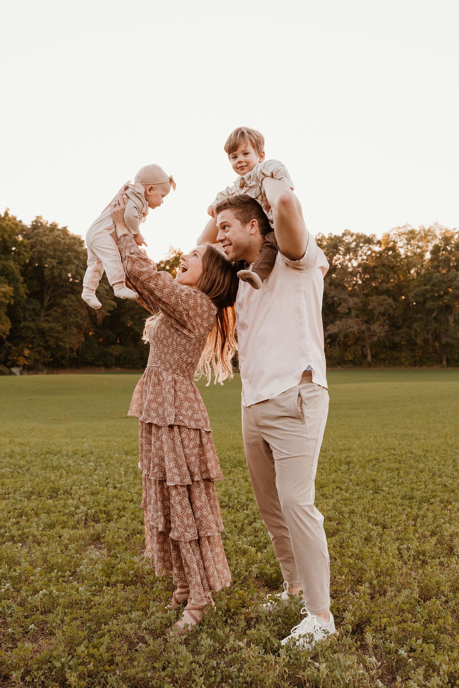 Happy family playing outdoors at sunset, parents lifting two young children in a green field with trees in background.