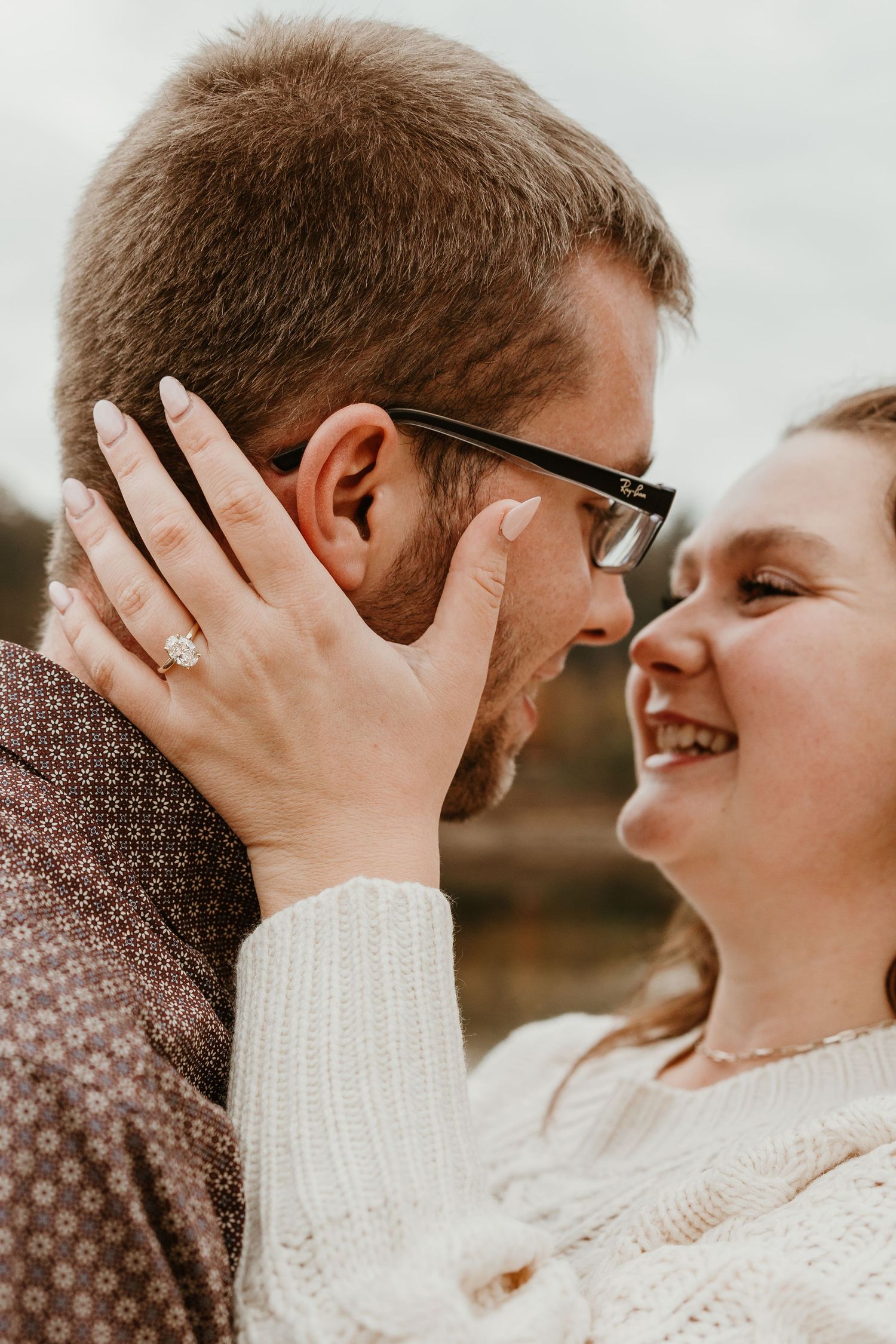 Close-up of smiling couple about to kiss, woman wearing engagement ring touching mans face, wearing glasses outdoors.