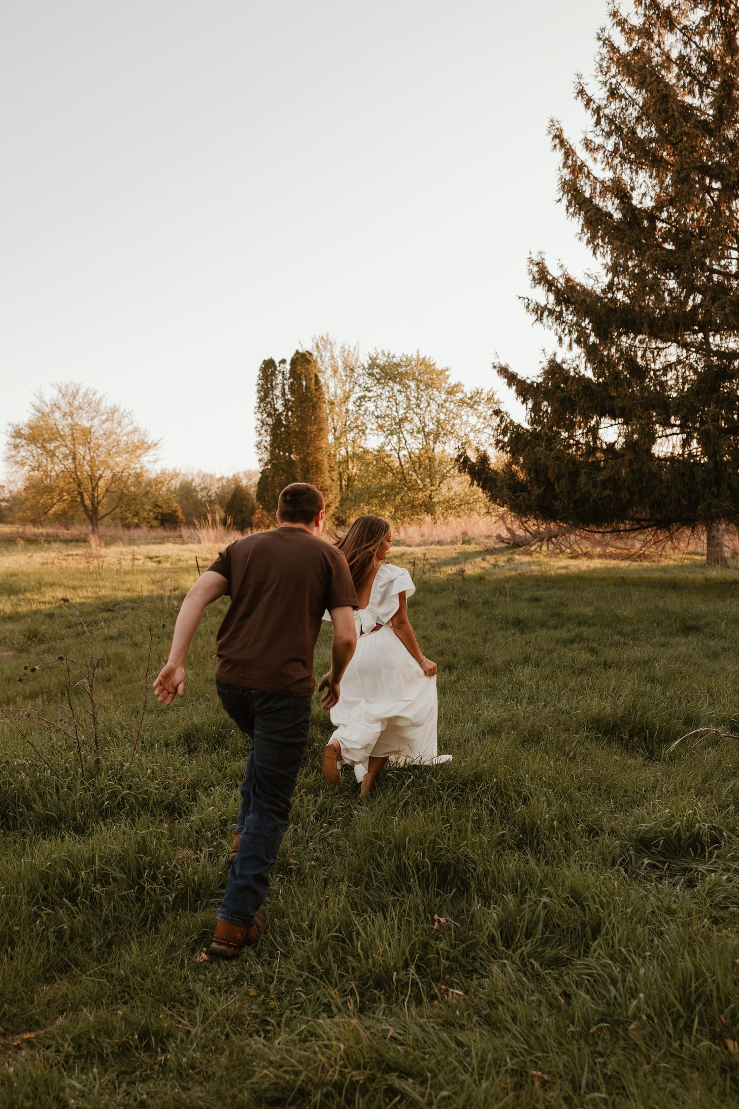 A couple runs hand-in-hand through a lush green field at golden hour, surrounded by trees in a romantic outdoor setting.