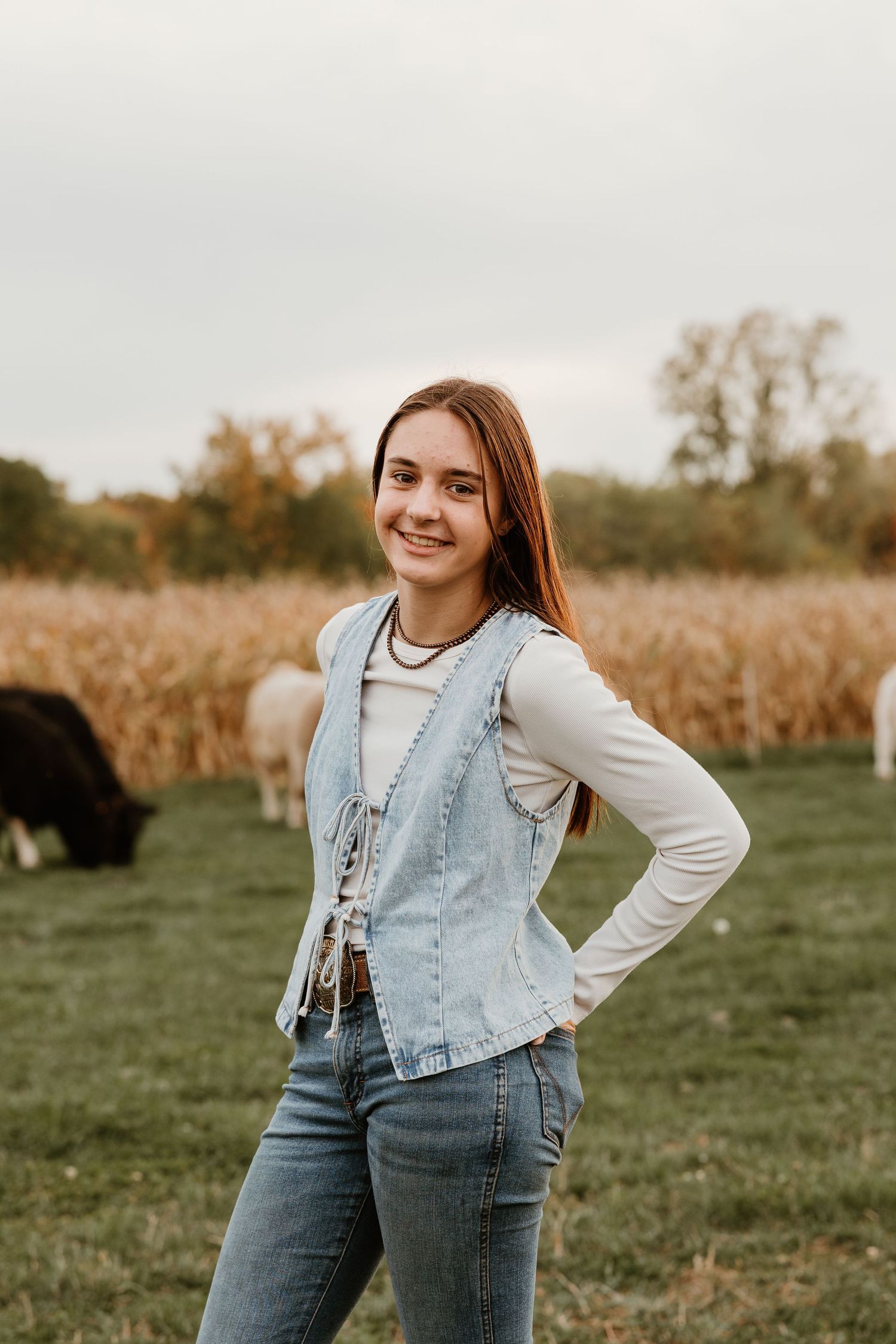 Smiling teenage girl in denim vest and jeans posing on a farm with cattle and cornfield in background.