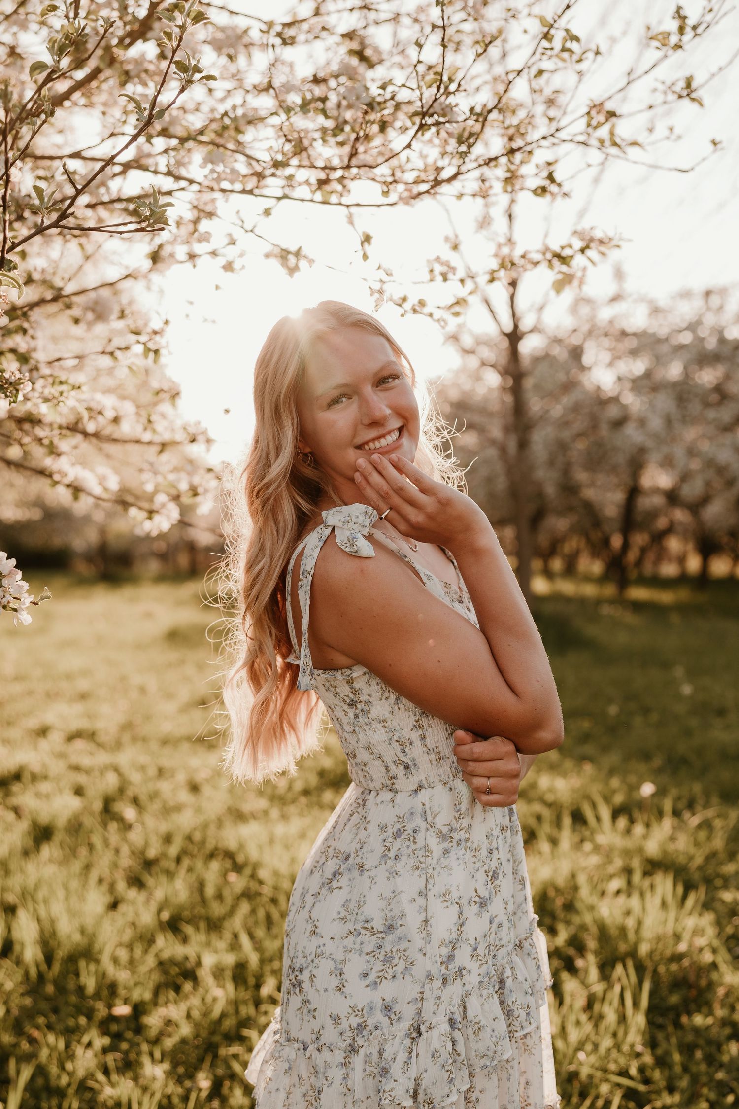 Young blonde woman in floral dress laughing in golden hour sunlight among blooming spring cherry blossom trees.