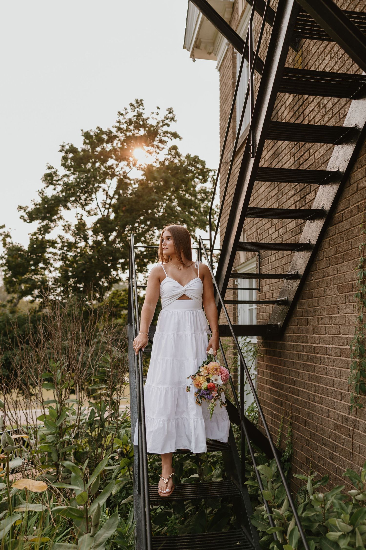 Woman in white midi dress holding colorful bouquet stands on metal fire escape stairs beside brick building at sunset.