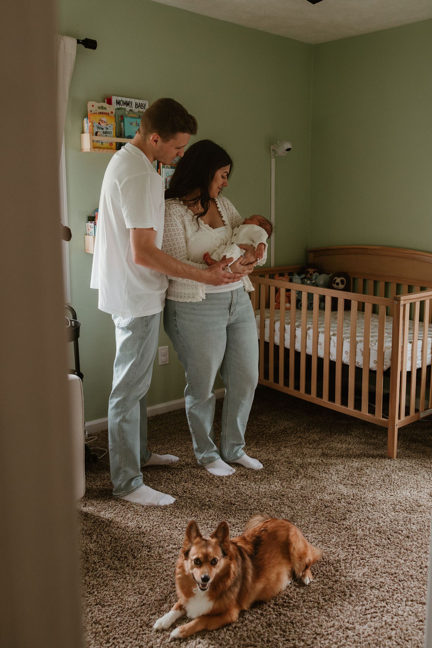 New parents holding newborn in green nursery with wooden crib, corgi dog lying on carpet nearby.